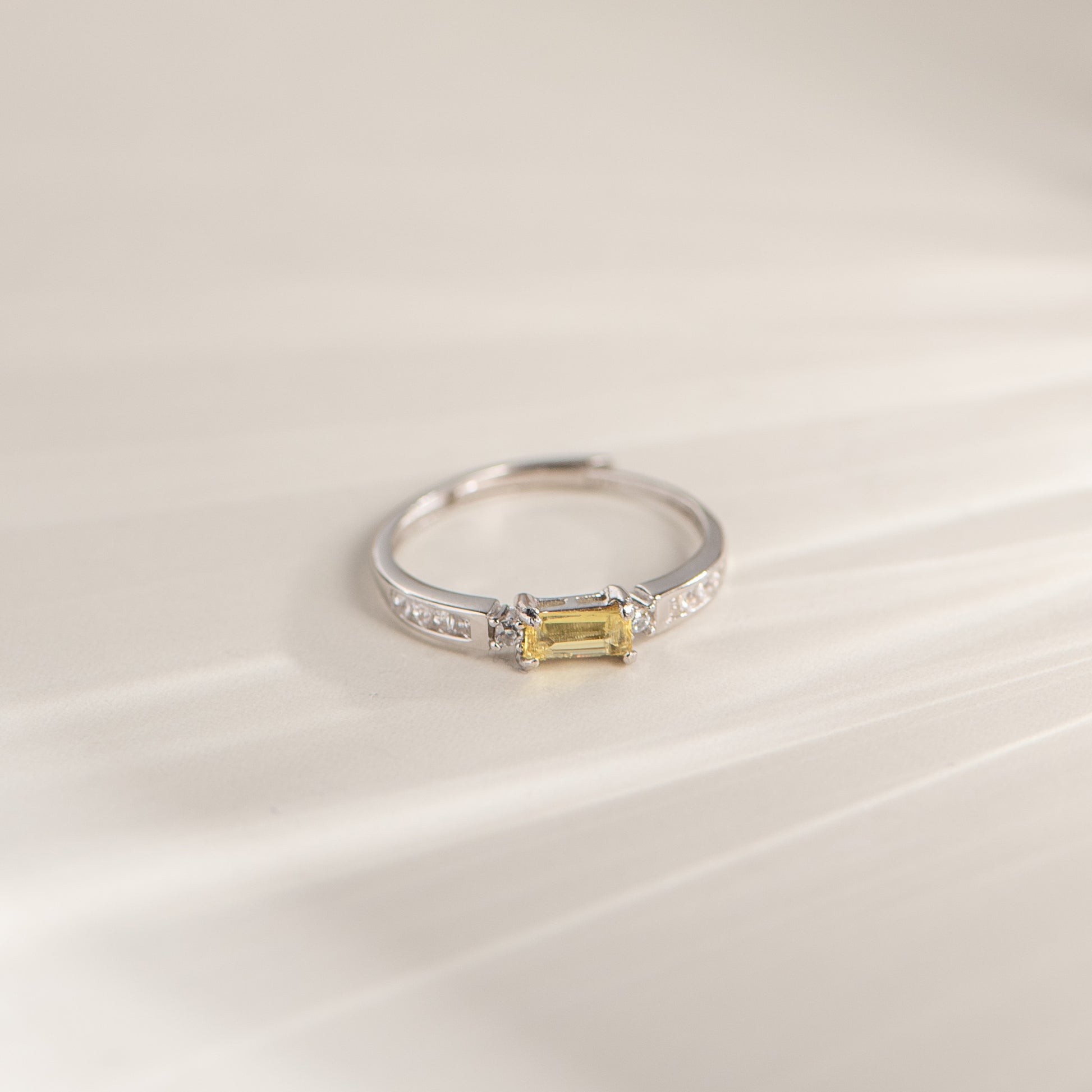 A silver ring featuring a rectangular yellow gemstone in a prong setting, accented with small clear stones along the band, photographed on a soft cream-colored surface.