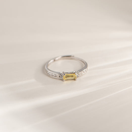 A silver ring featuring a rectangular yellow gemstone in a prong setting, accented with small clear stones along the band, photographed on a soft cream-colored surface.