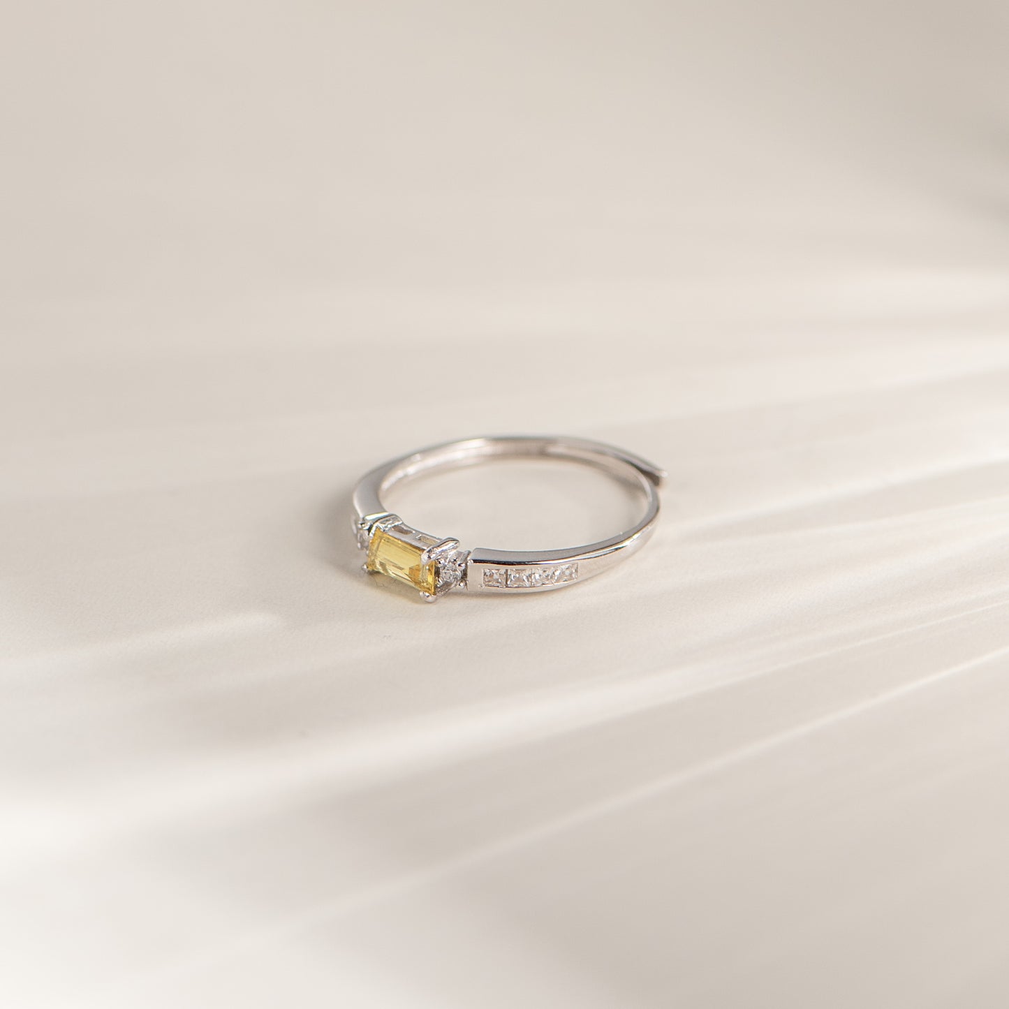 A silver ring with a small rectangular yellow gemstone, photographed on a soft cream-colored surface with a shallow depth of field.