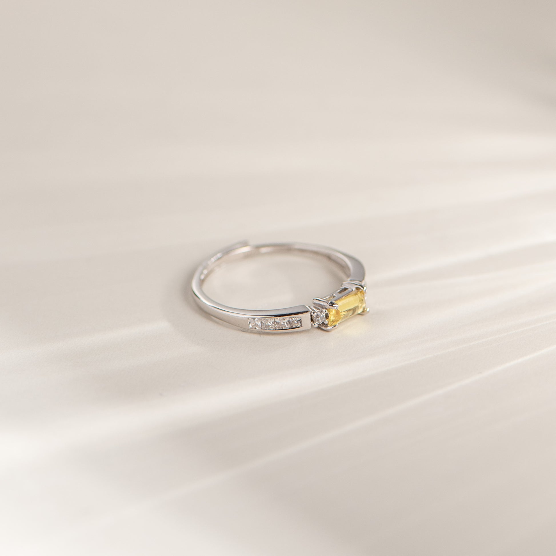 A silver ring featuring a small rectangular yellow gemstone and a row of tiny clear stones on the band, photographed on a soft cream-colored surface.
