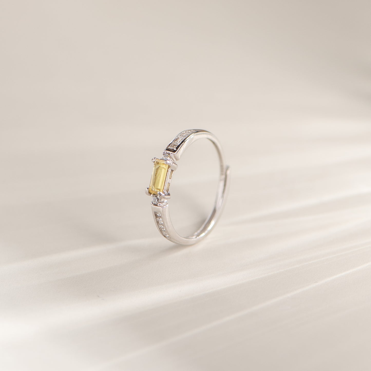 A silver-toned open band ring featuring a rectangular yellow gemstone in a prong setting, accented with small clear stones on the band, photographed on a soft light beige background.