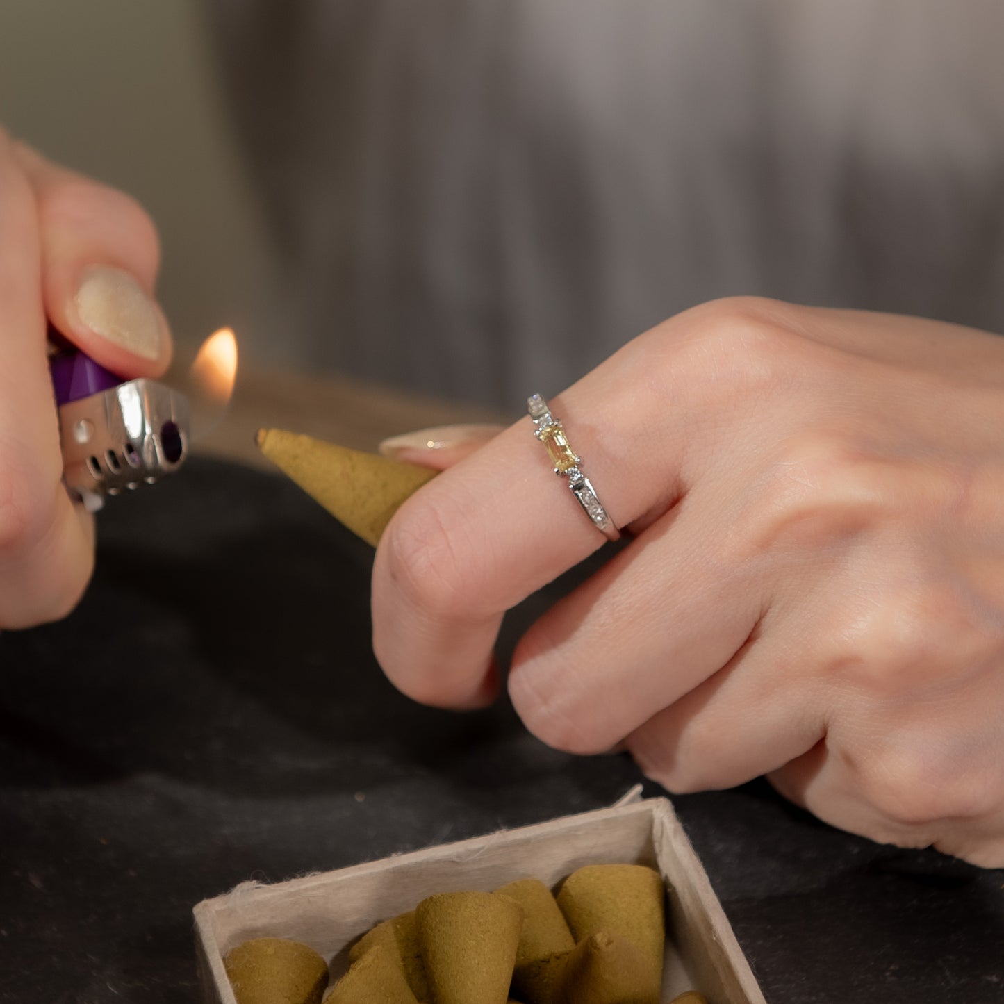 A close-up lifestyle shot of a person's hands, with one hand wearing a delicate silver ring and the other holding a cone of incense while lighting it with a purple and silver lighter. A small box of incense cones is visible in the foreground.