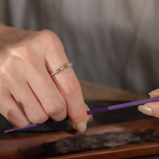 A close-up lifestyle shot of a hand wearing a silver band ring with rectangular and round-cut clear stones, holding a purple incense stick over a burning incense burner.