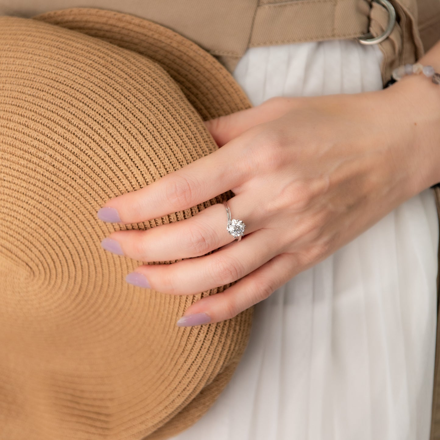 Hand wearing a diamond ring with a straw hat and white fabric in the background