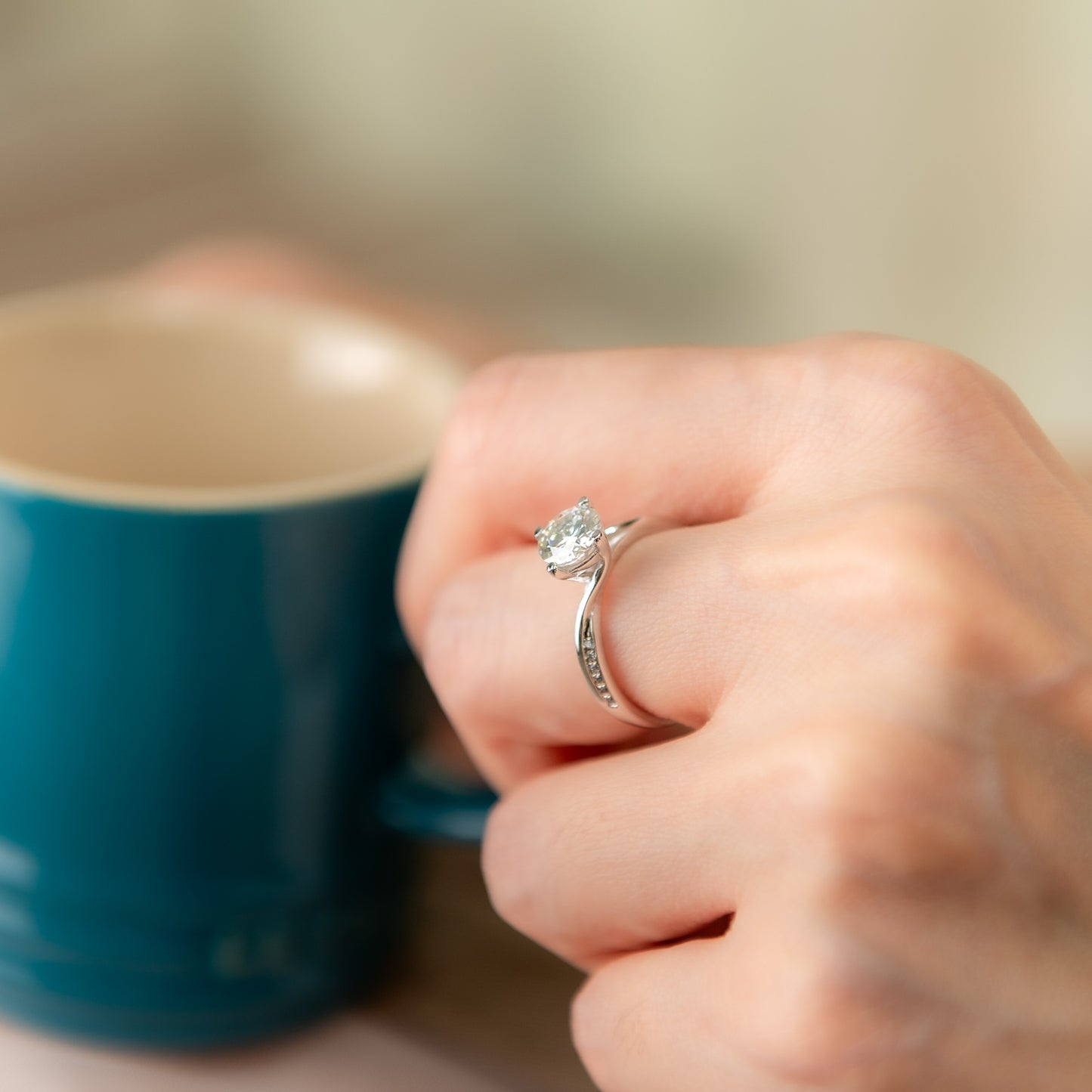 Hand wearing a diamond ring with a blurred blue mug in the background