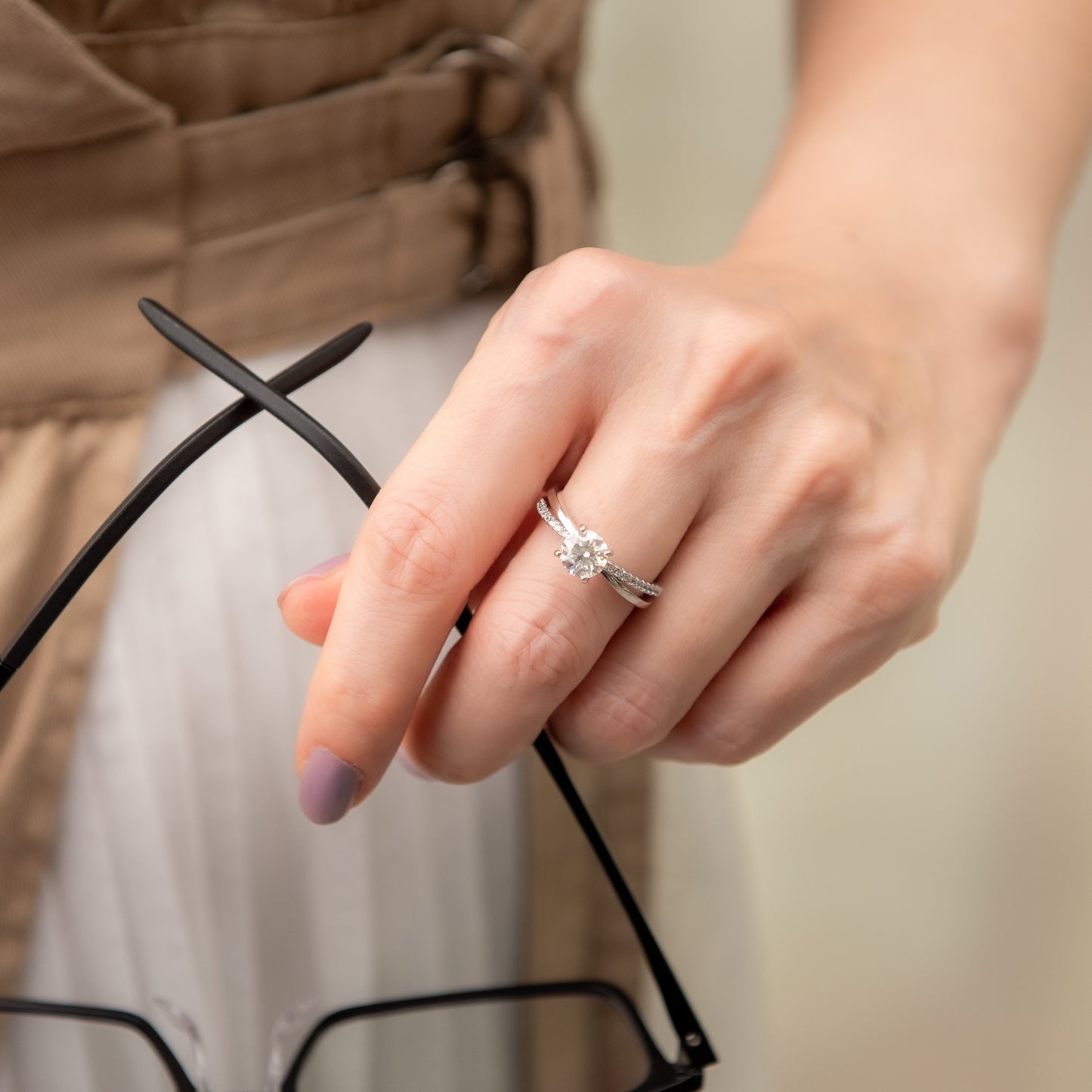 Hand wearing a silver ring with a diamond, holding eyeglasses against a neutral background