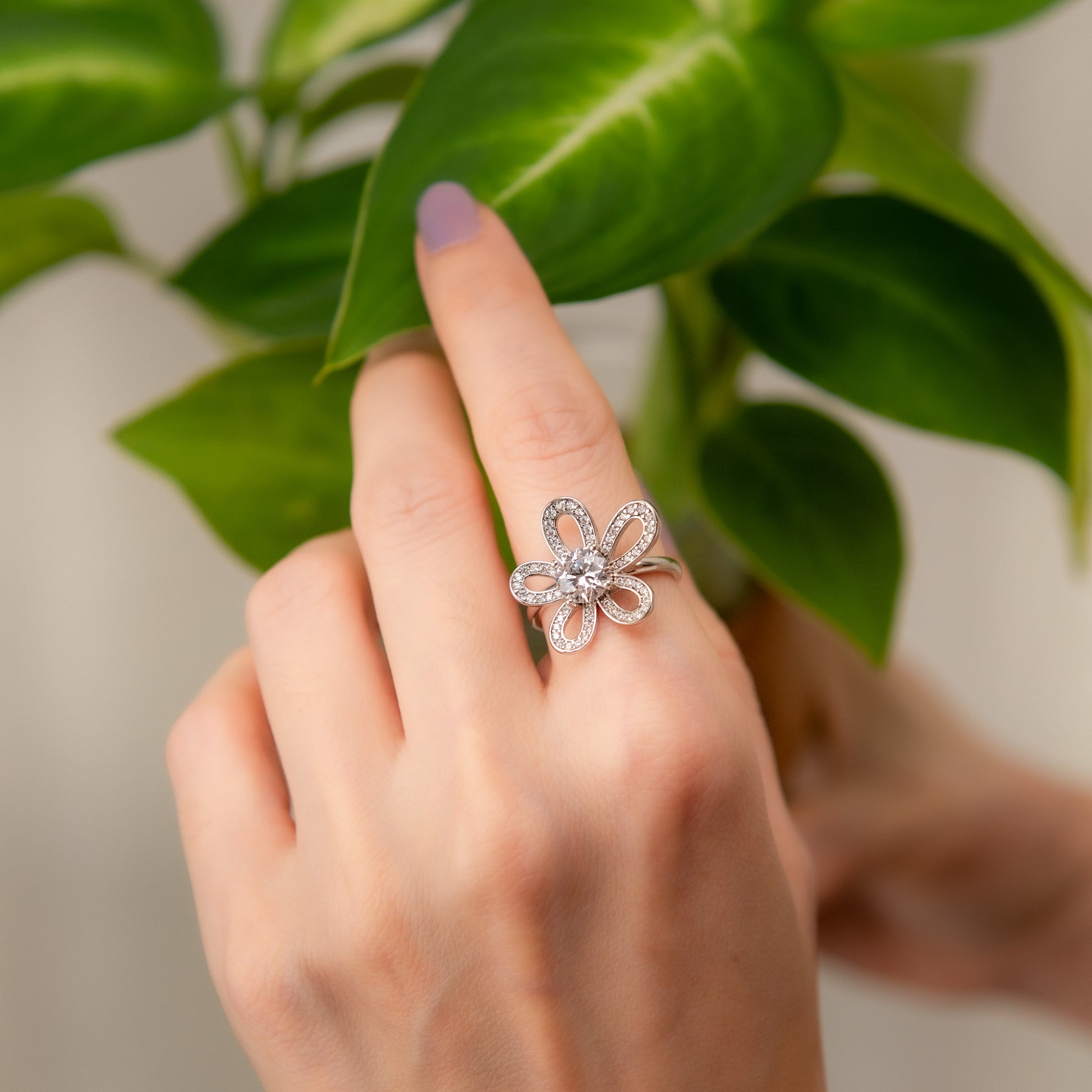 Hand wearing a silver flower-shaped ring with green leaves in the background