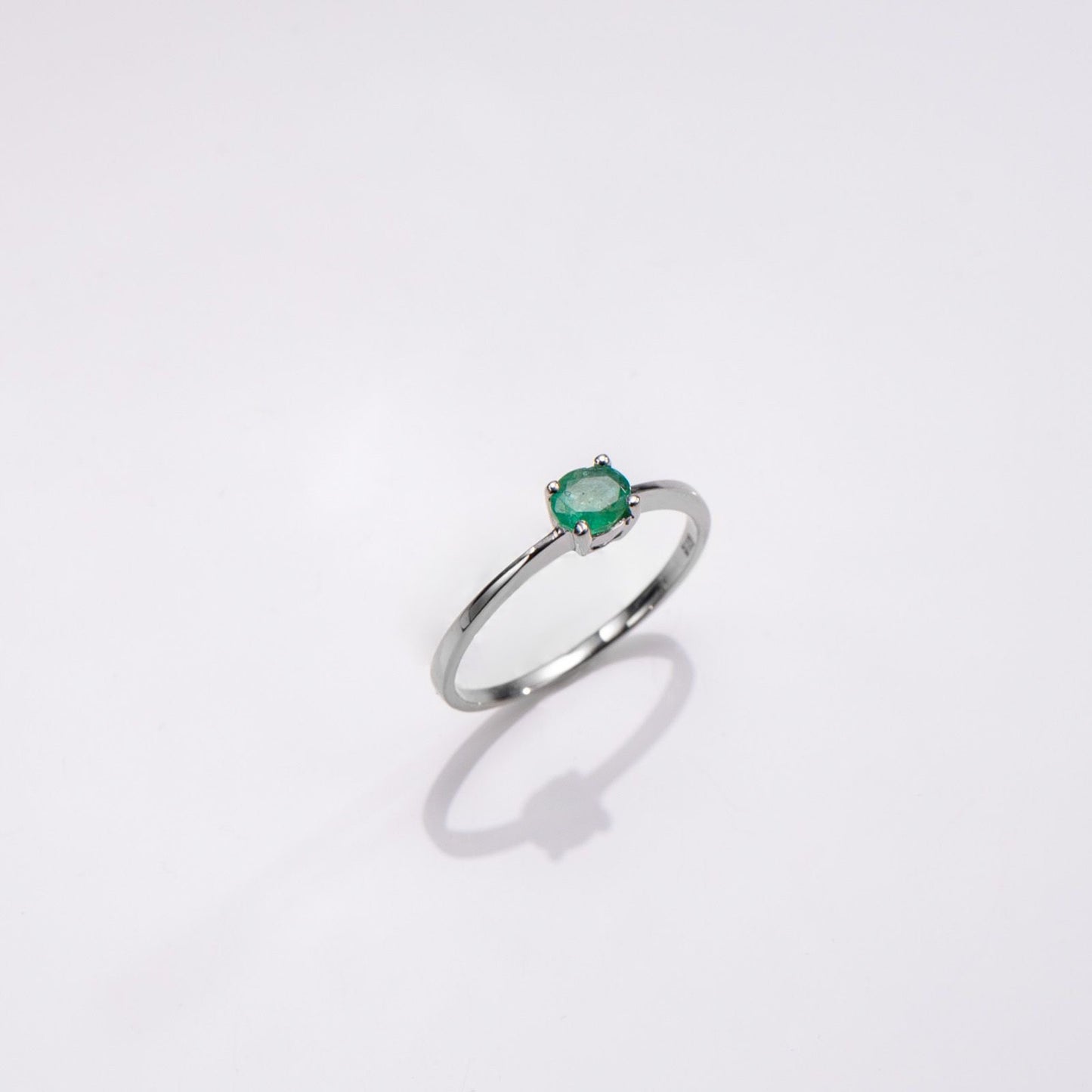 A silver ring with a small oval green gemstone in a prong setting, photographed on a clean white background with a soft shadow below.