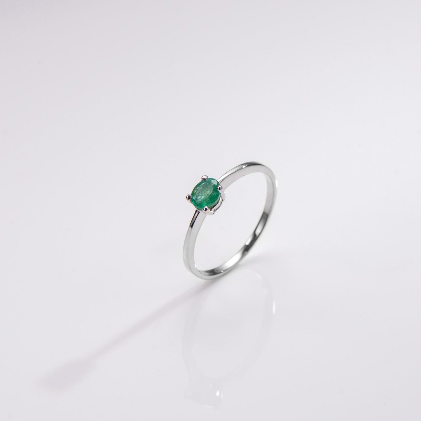 A silver ring with a small oval green gemstone in a prong setting, photographed on a clean white reflective surface.
