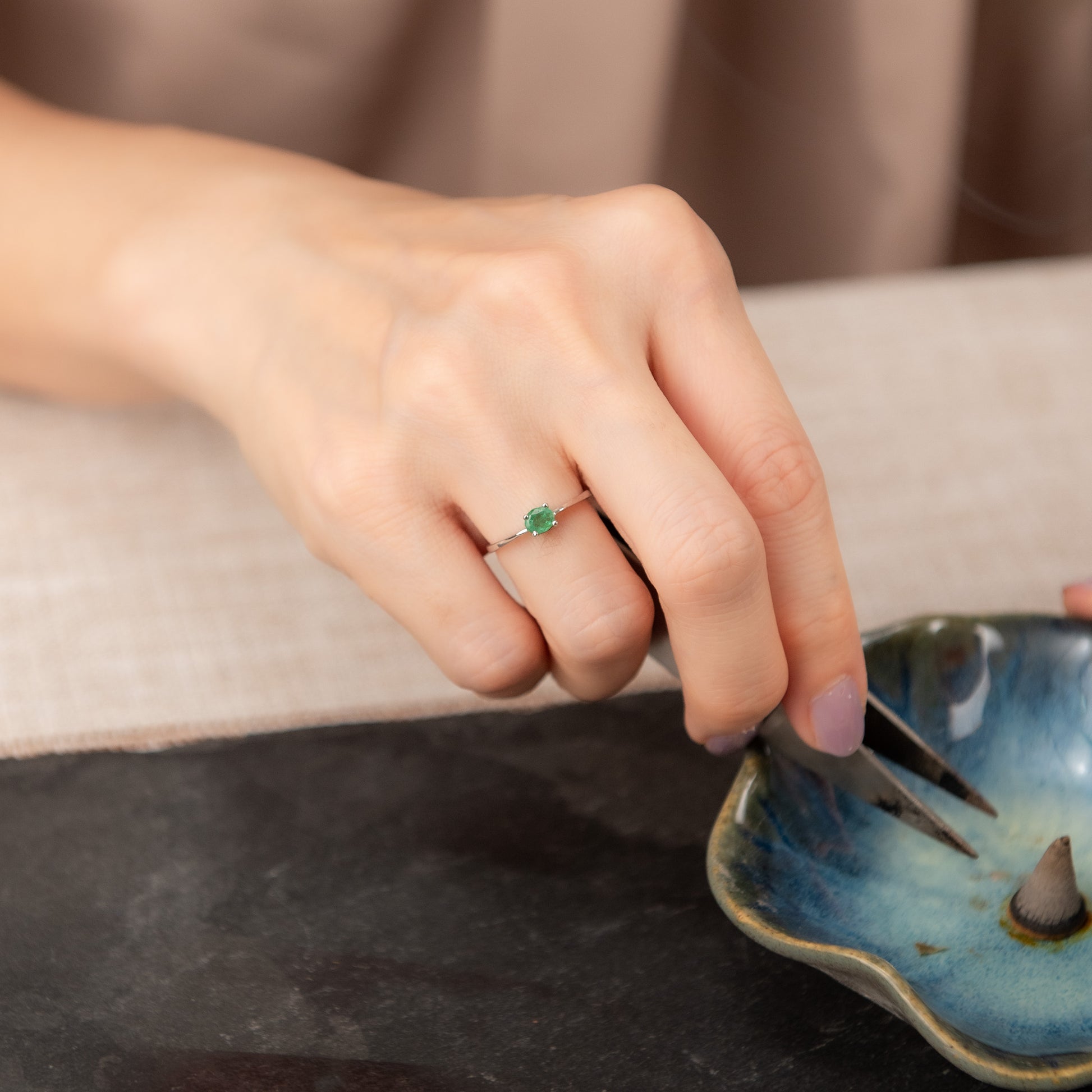 A close-up of a hand wearing a delicate silver ring with a small green heart-shaped gemstone, while holding tweezers over a blue ceramic dish.
