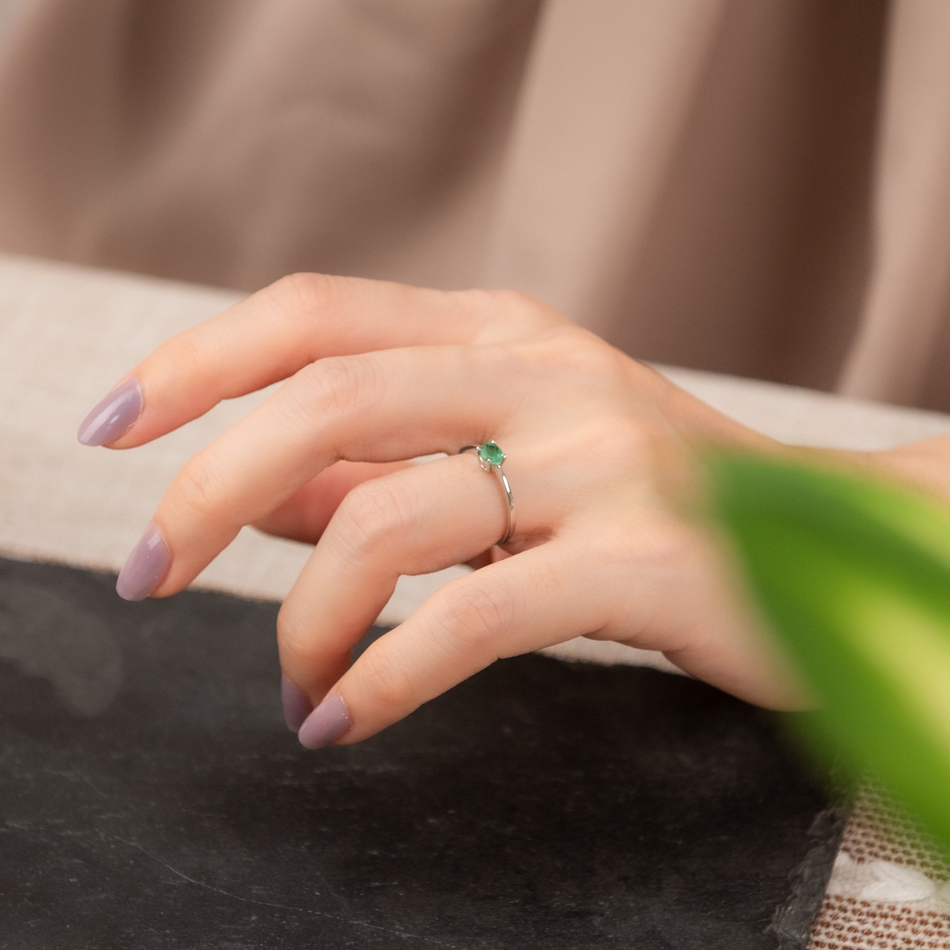 A close-up of a person's hand with lavender-painted nails resting on a dark surface, wearing a thin silver ring with a small green gemstone.