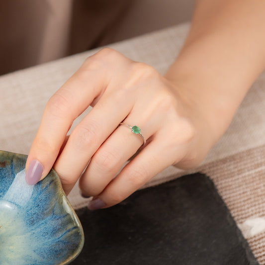 A close-up of a person's hand wearing a delicate silver ring with a small green gemstone, resting near a blue marbled bowl on a neutral textured surface.