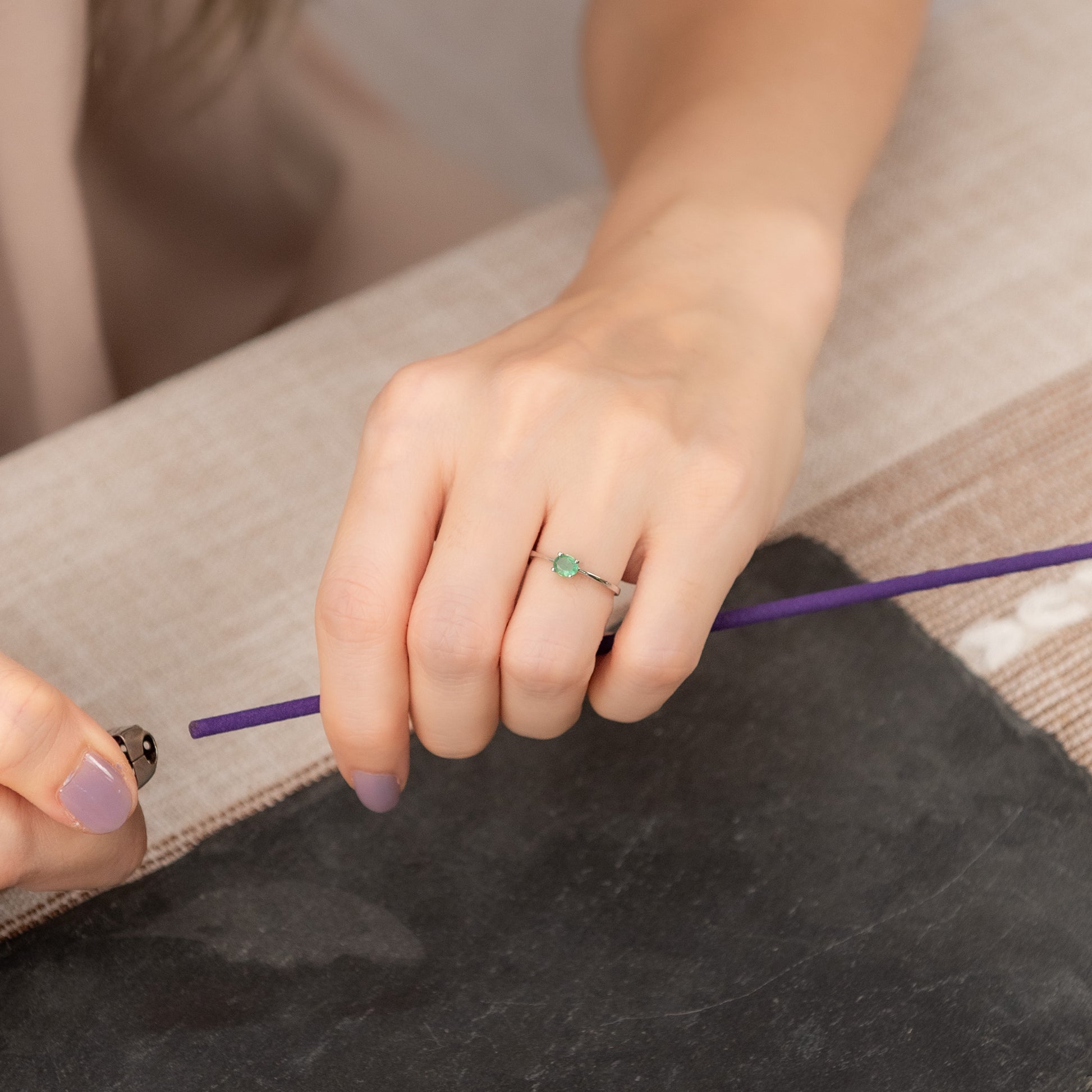 A close-up of a person's hands holding a purple incense stick, wearing a delicate silver ring with a small green stone on one finger. The background features a textured beige woven mat and a dark charcoal slate surface.