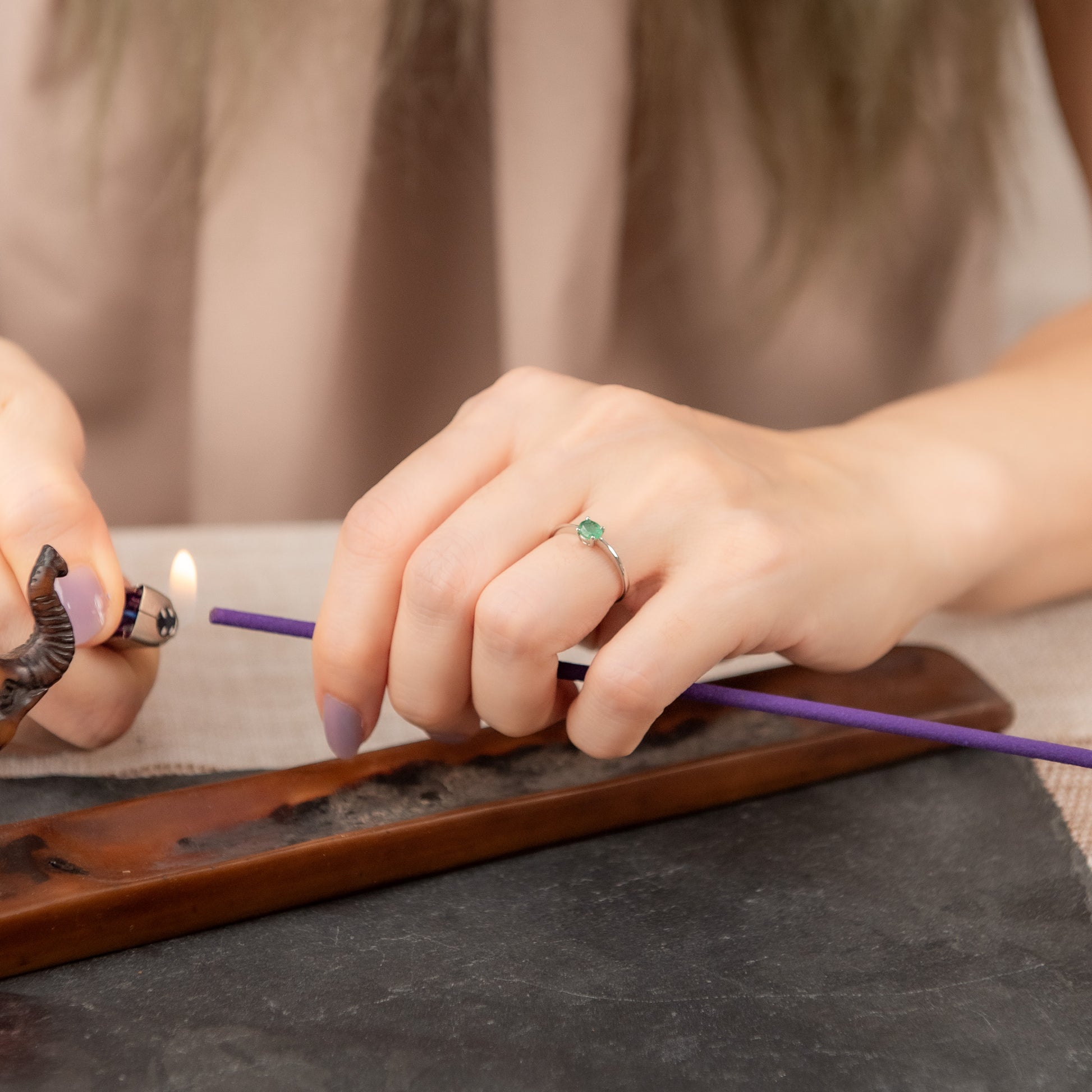 A close-up lifestyle shot of a person's hands lighting a purple incense stick over a wooden tray, with a silver ring featuring a green gemstone worn on one finger.