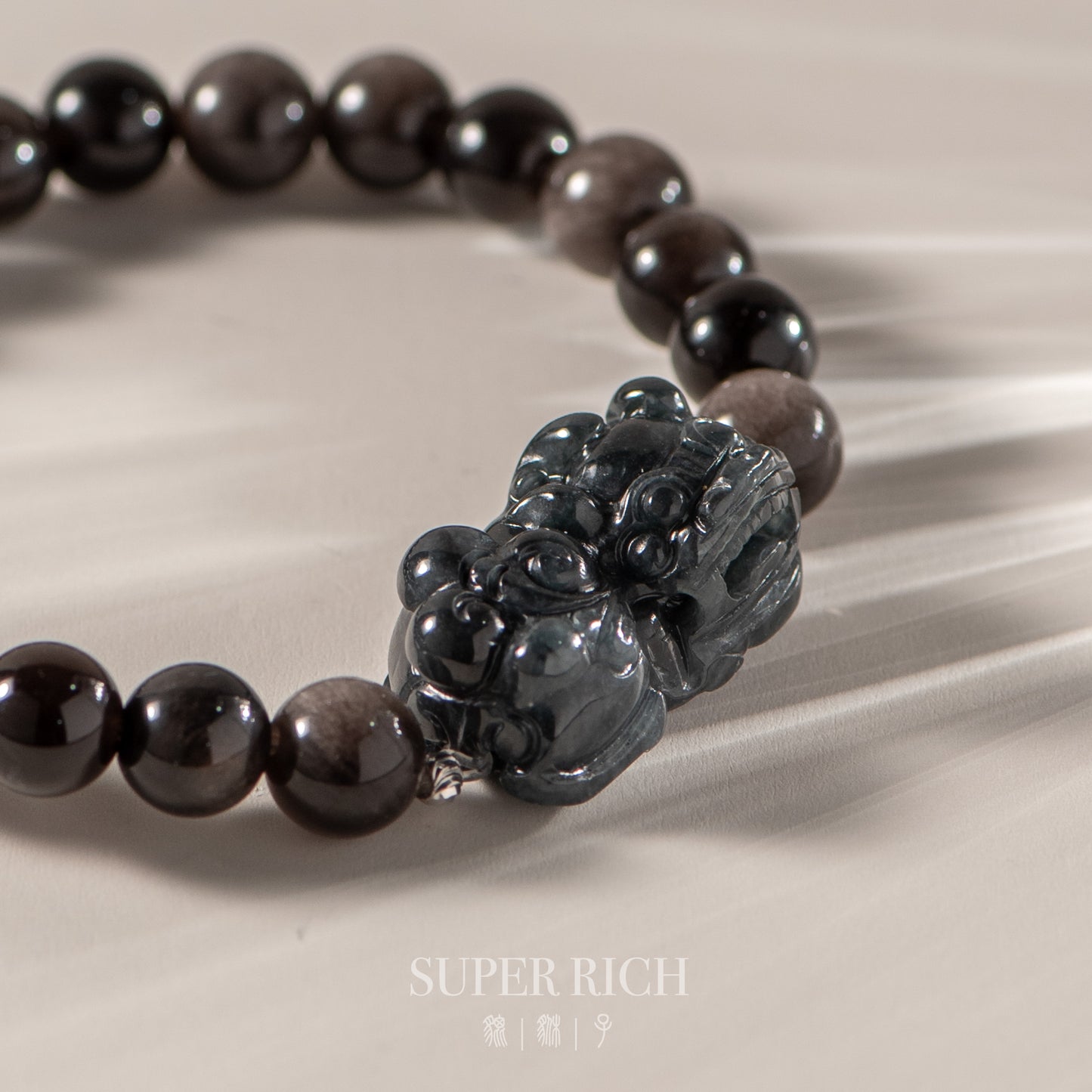Black beaded bracelet with a carved pendant on a beige fabric background, featuring the brand 'Super Rich'.