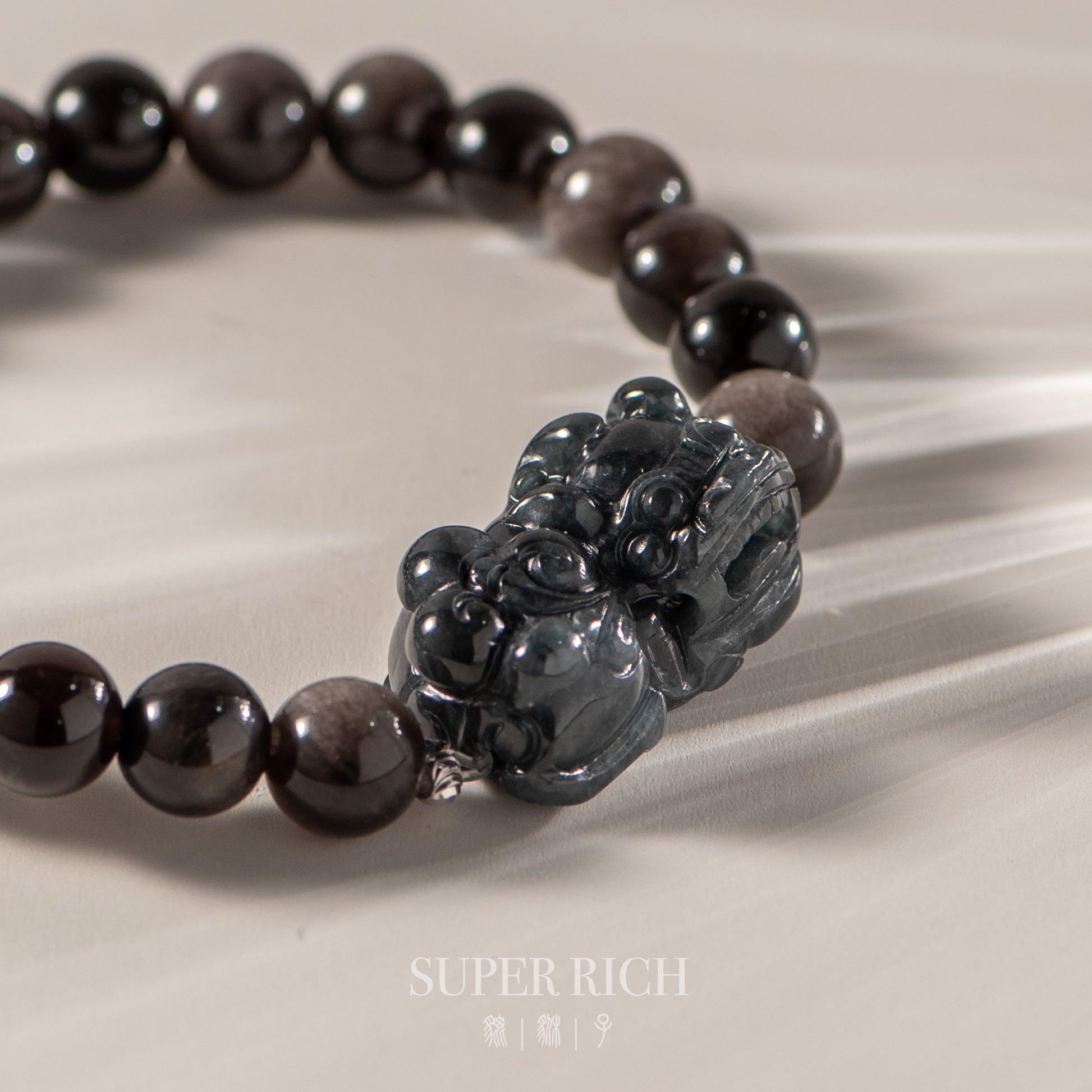 Black beaded bracelet with a carved pendant on a beige fabric background, featuring the brand 'Super Rich'.