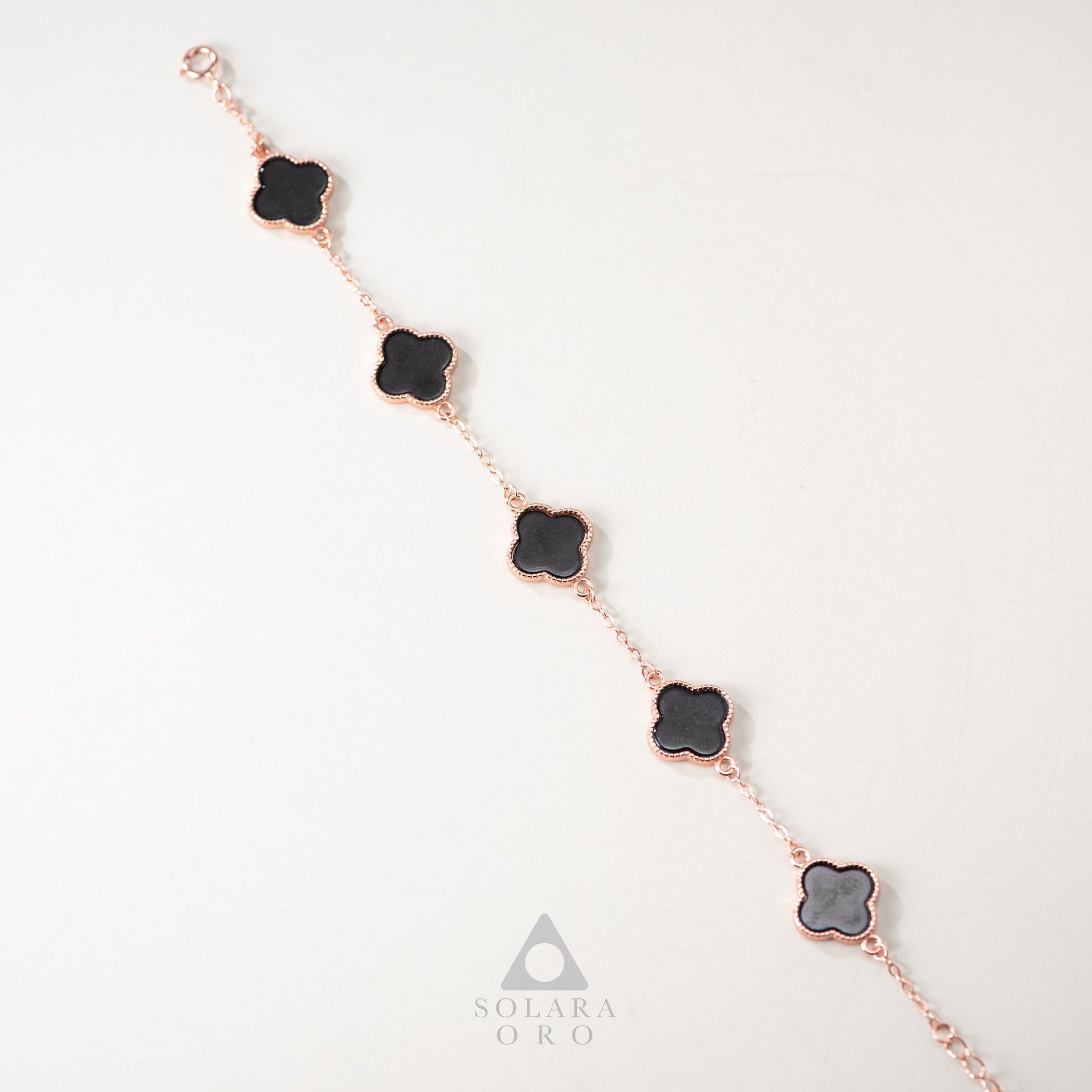 Rose gold bracelet with black clover-shaped charms on a light gray background, featuring the Solara Oro brand.
