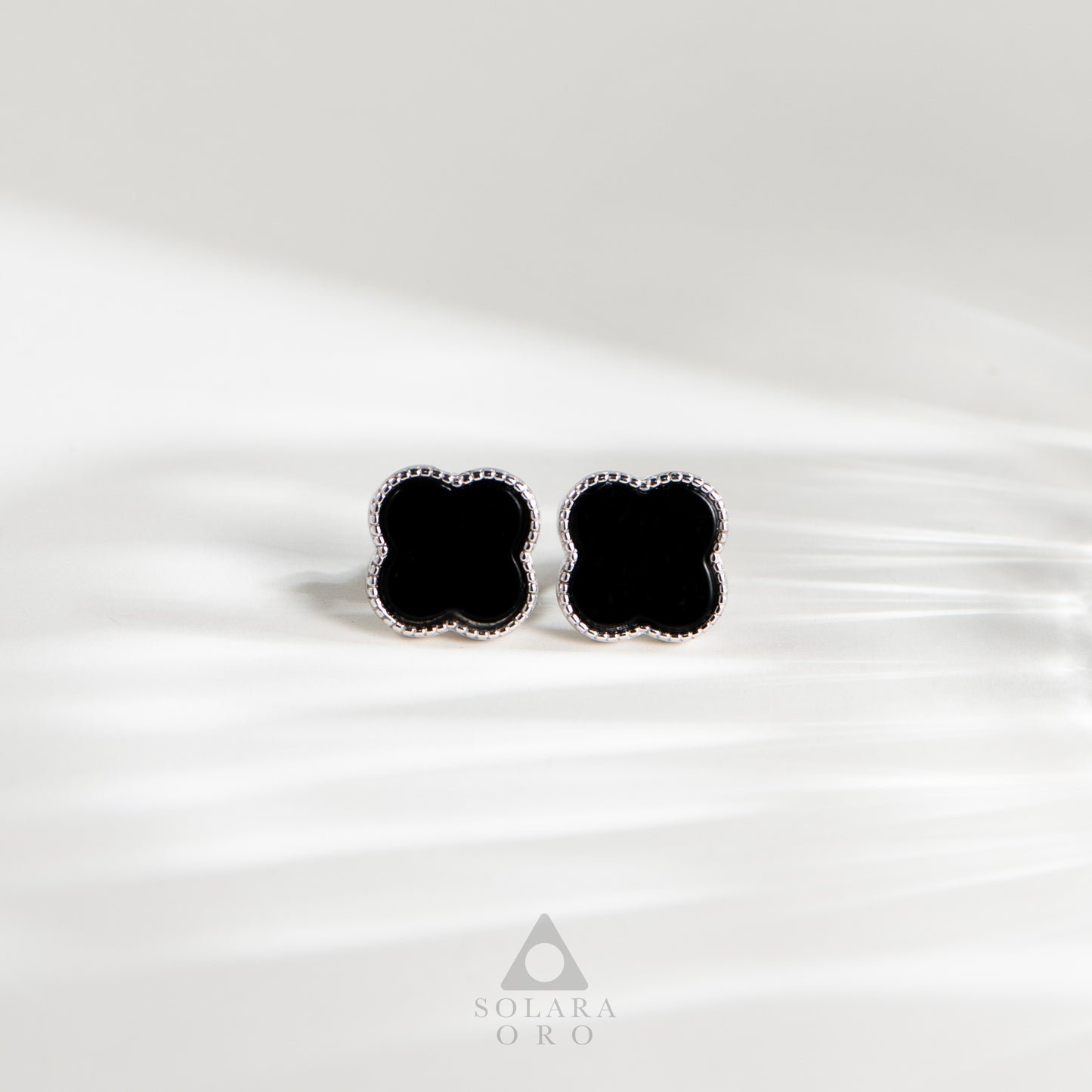 Black clover-shaped earrings on a white satin background with Solara Oro logo.