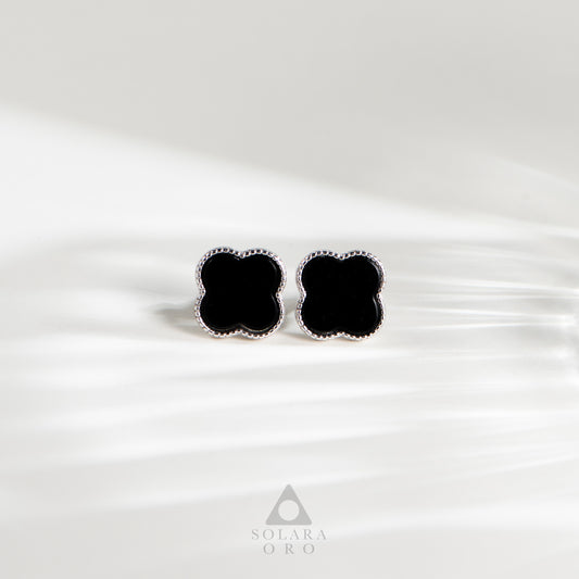 Black clover-shaped earrings on a white satin background with Solara Oro logo.