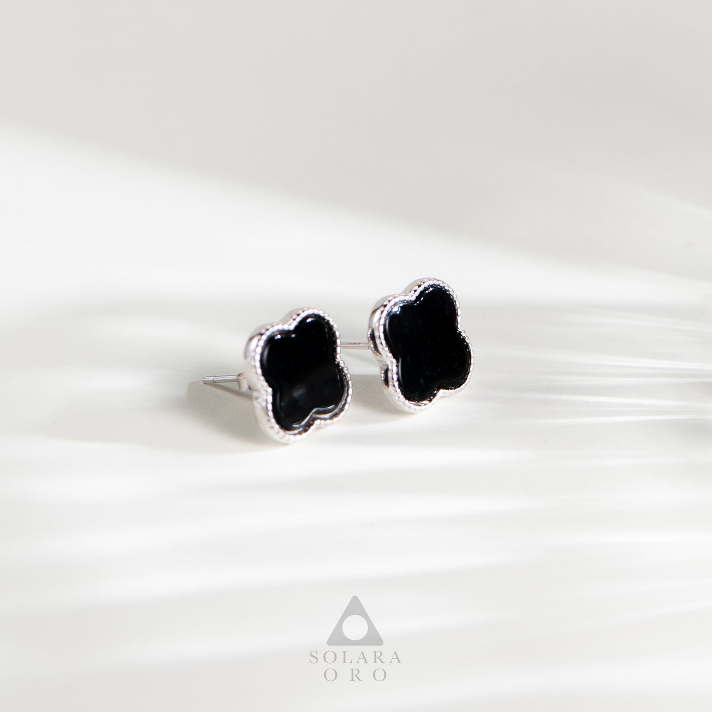 Black clover-shaped earrings on a white background with Solara Oro logo.