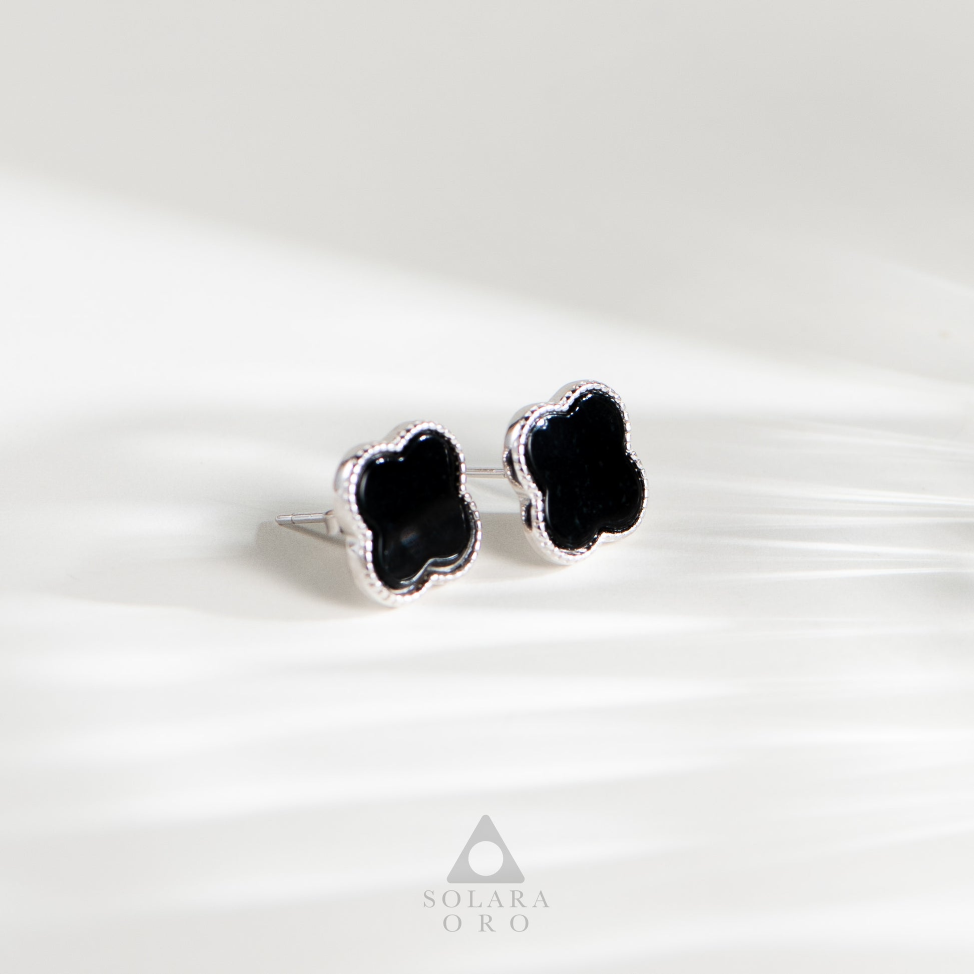 Black clover-shaped earrings on a white background with Solara Oro logo.