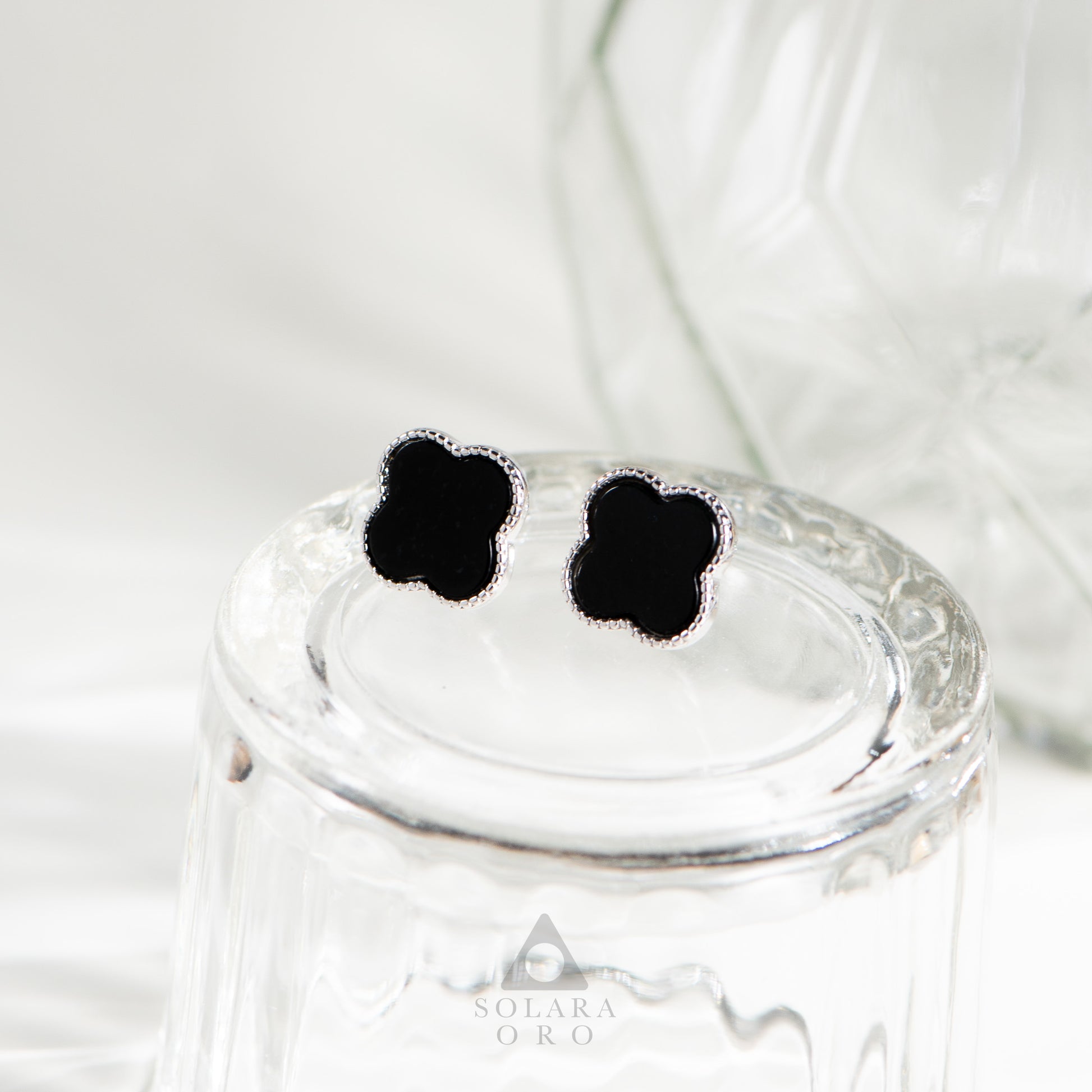 Black flower-shaped earrings on a glass jar with 'Solerara Oro' branding.