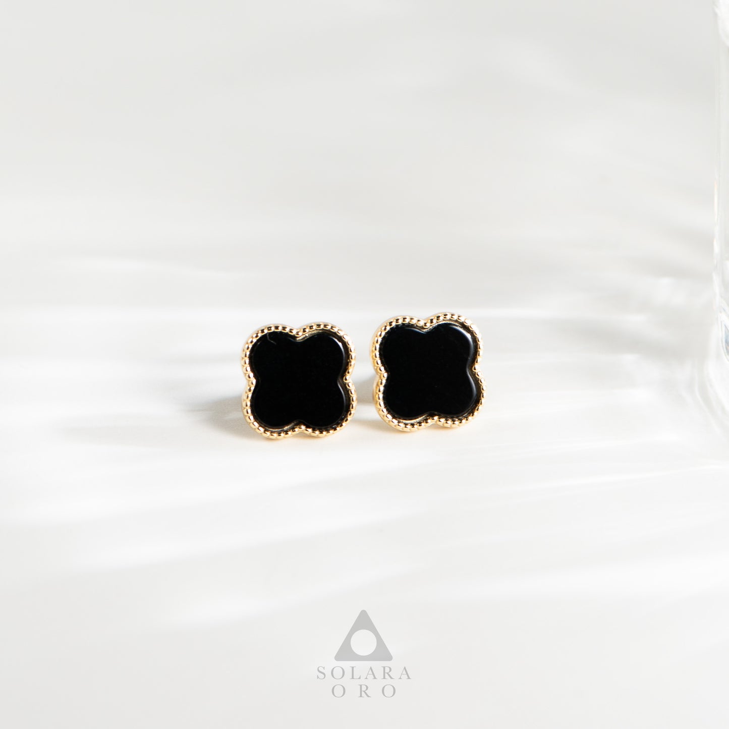 Black clover-shaped earrings with gold accents on a white background, branded 'Soleara Oro'.