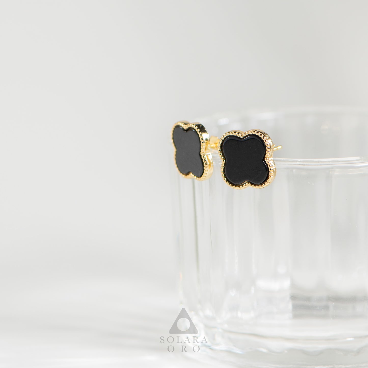 Black clover-shaped earrings with gold accents on a white background, branded 'Solaris Oro'.