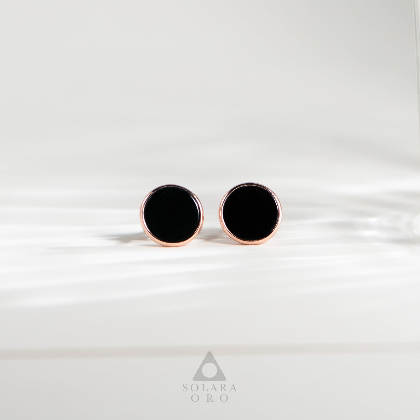 Black round earrings with rose gold posts on a white background, branded 'Solara Oro'.