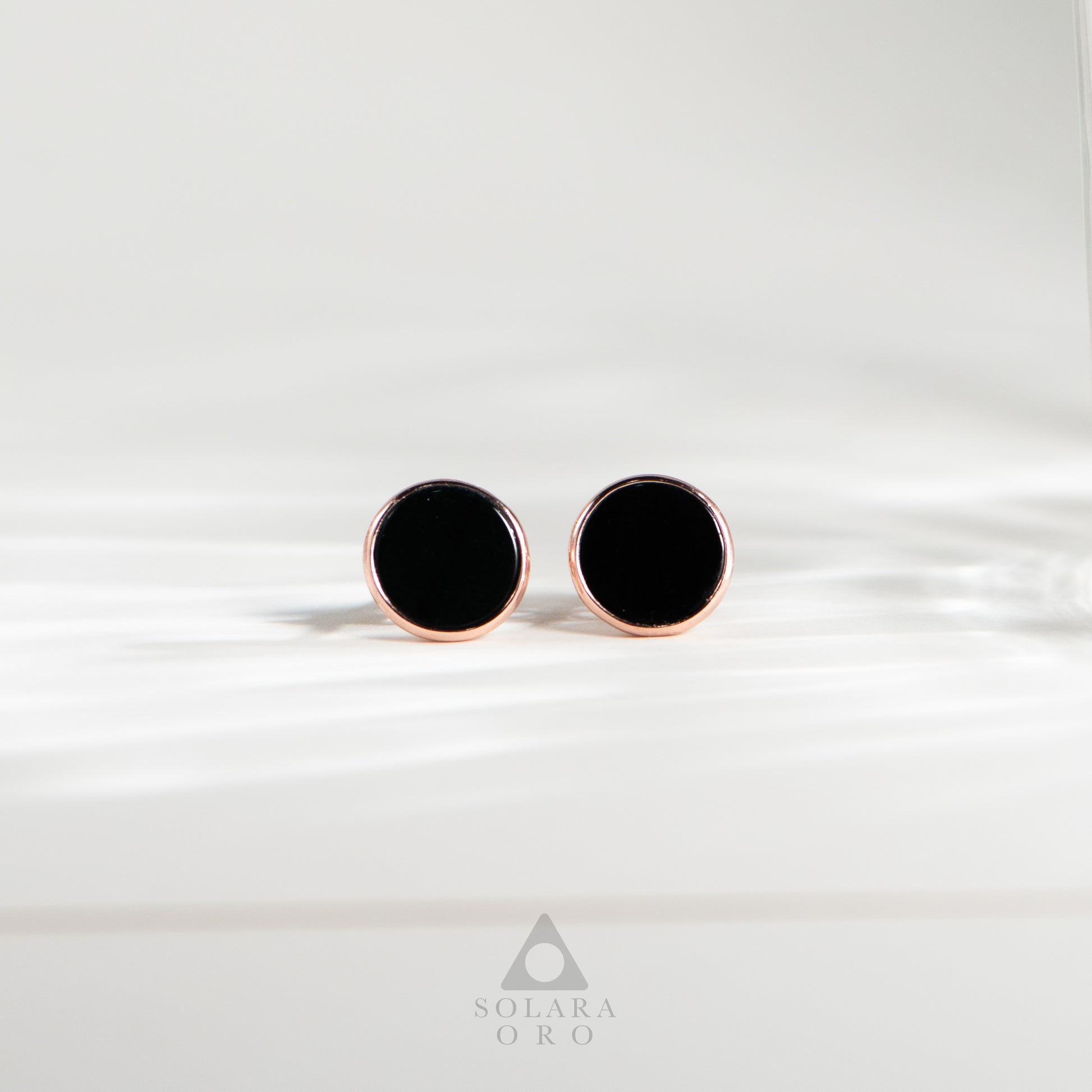Black round earrings with rose gold posts on a white background, branded 'Solara Oro'.