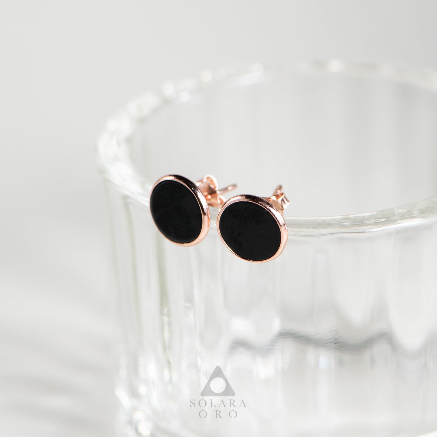 Black round earrings with rose gold accents on a glass surface, branded 'Solara Oro'.