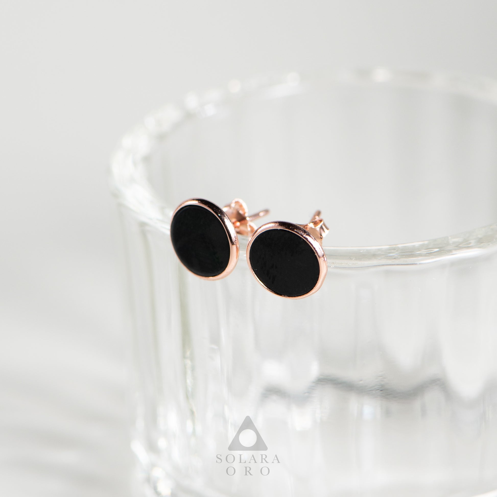 Black round earrings with rose gold accents on a glass surface, branded 'Solara Oro'.