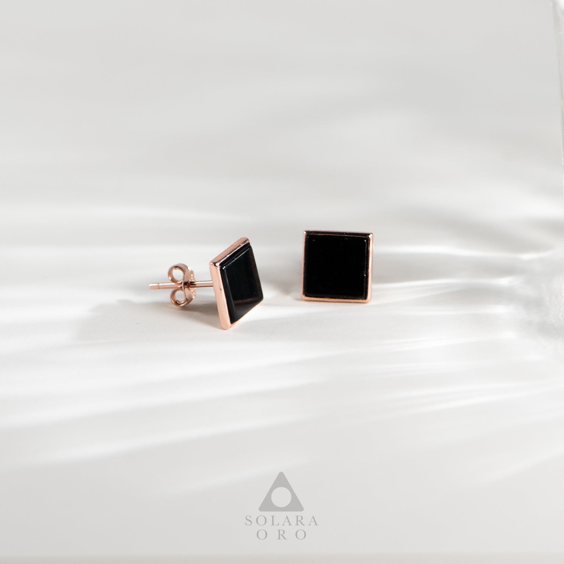 Black square stud earrings with rose gold posts on a white satin background, branded 'Solara Oro'.
