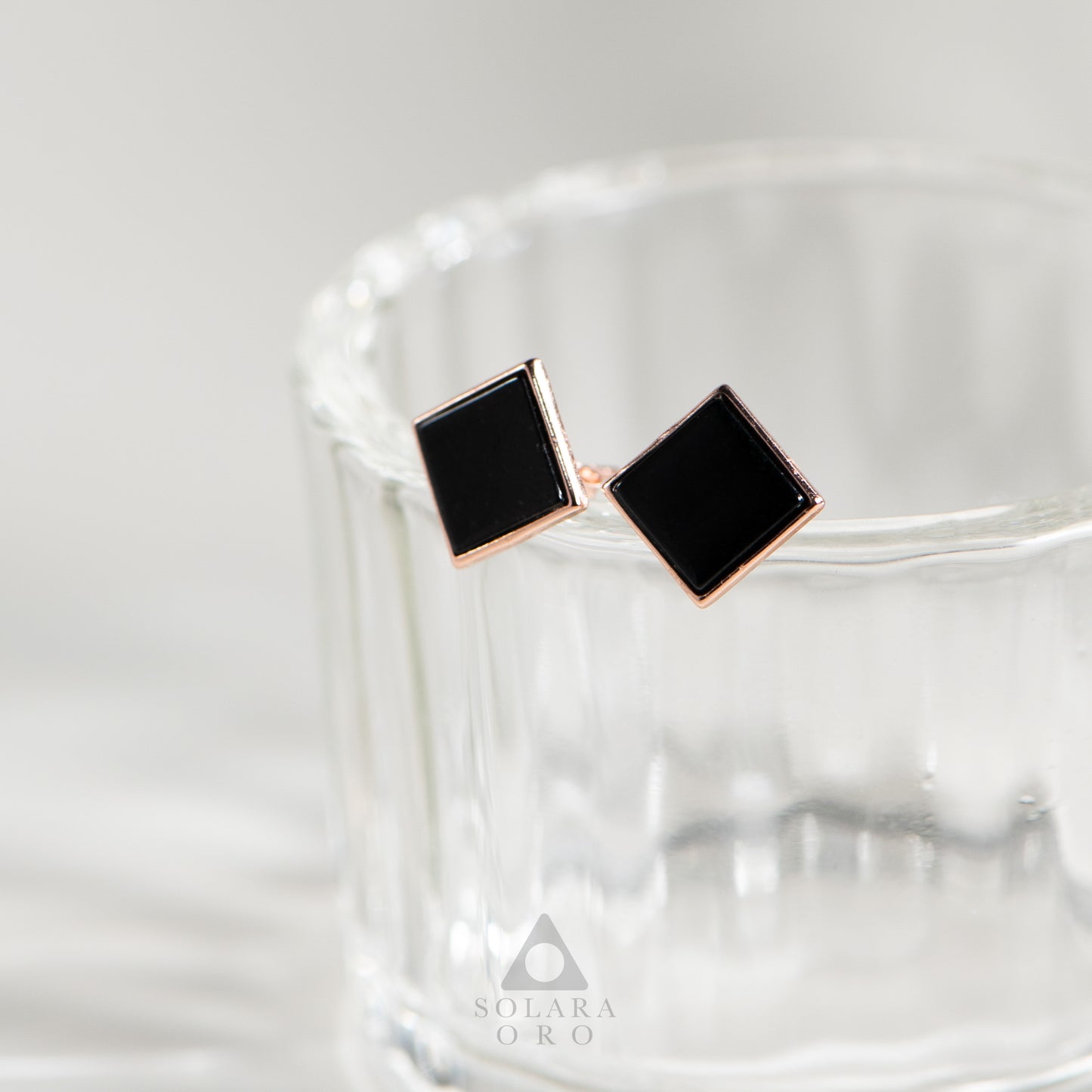 Black square stud earrings with rose gold accents on a white background, featuring the Solara Oro brand.