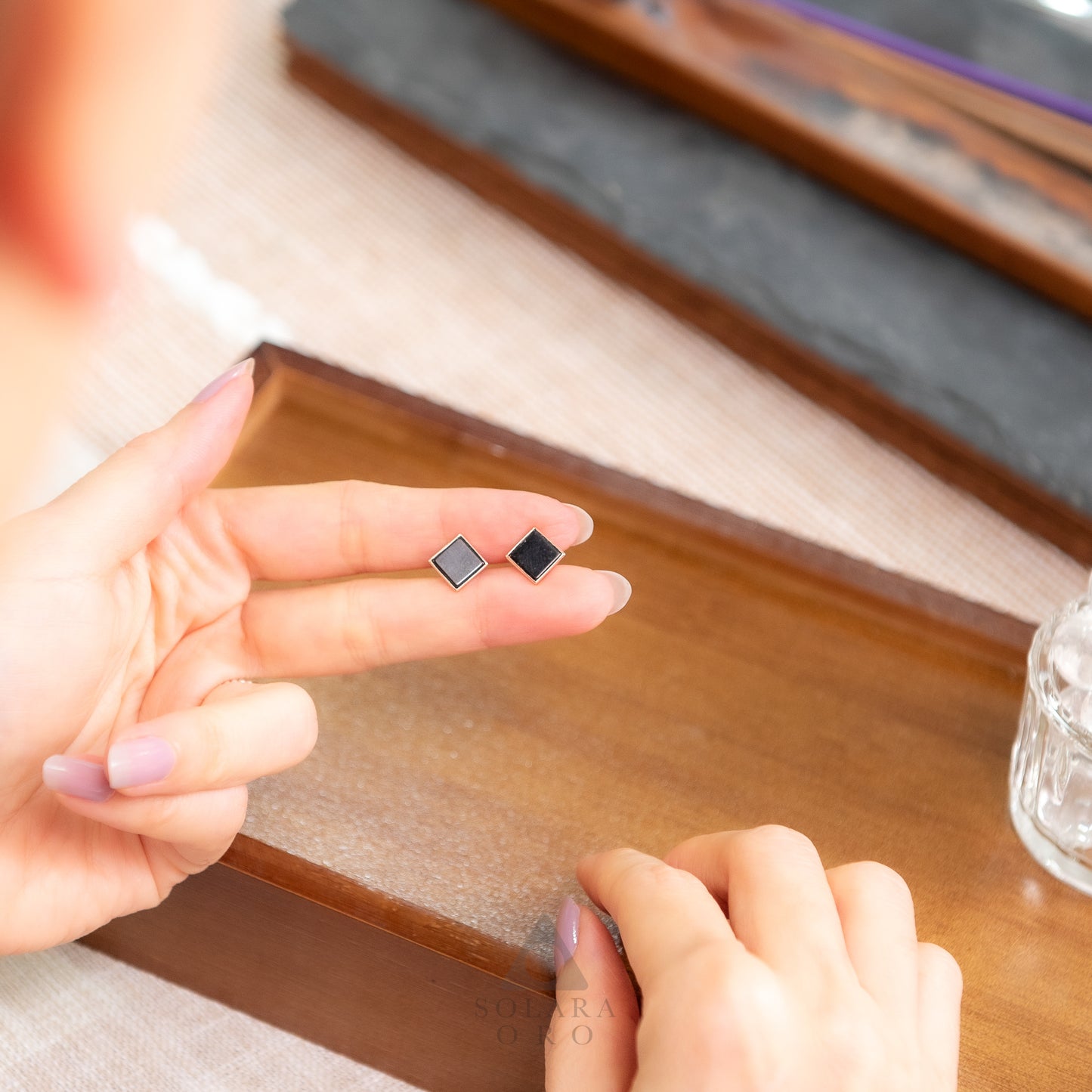 Person holding two black square earrings on a wooden surface