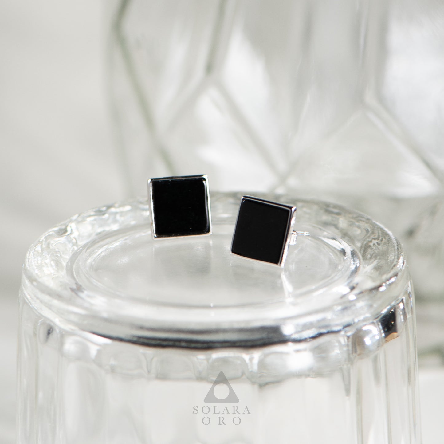 Black square stud earrings on a clear glass stand with 'Solara Oro' branding.