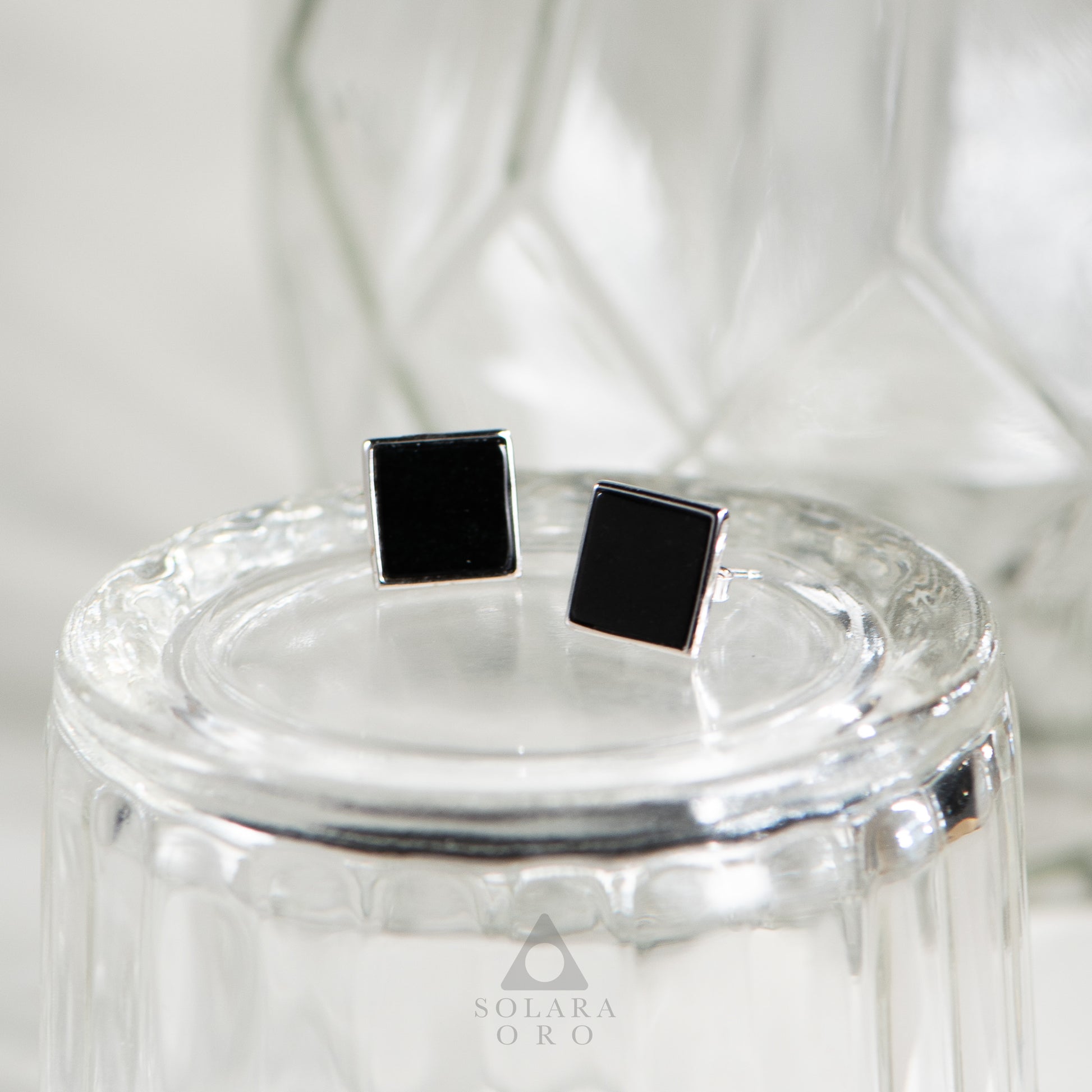 Black square stud earrings on a clear glass stand with 'Solara Oro' branding.