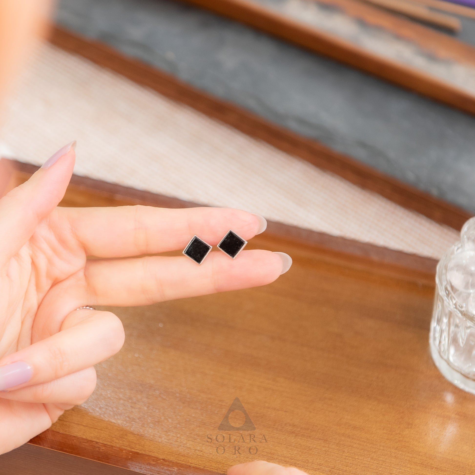 Person holding black square earrings on a wooden surface with 'Sulara Doro' branding.