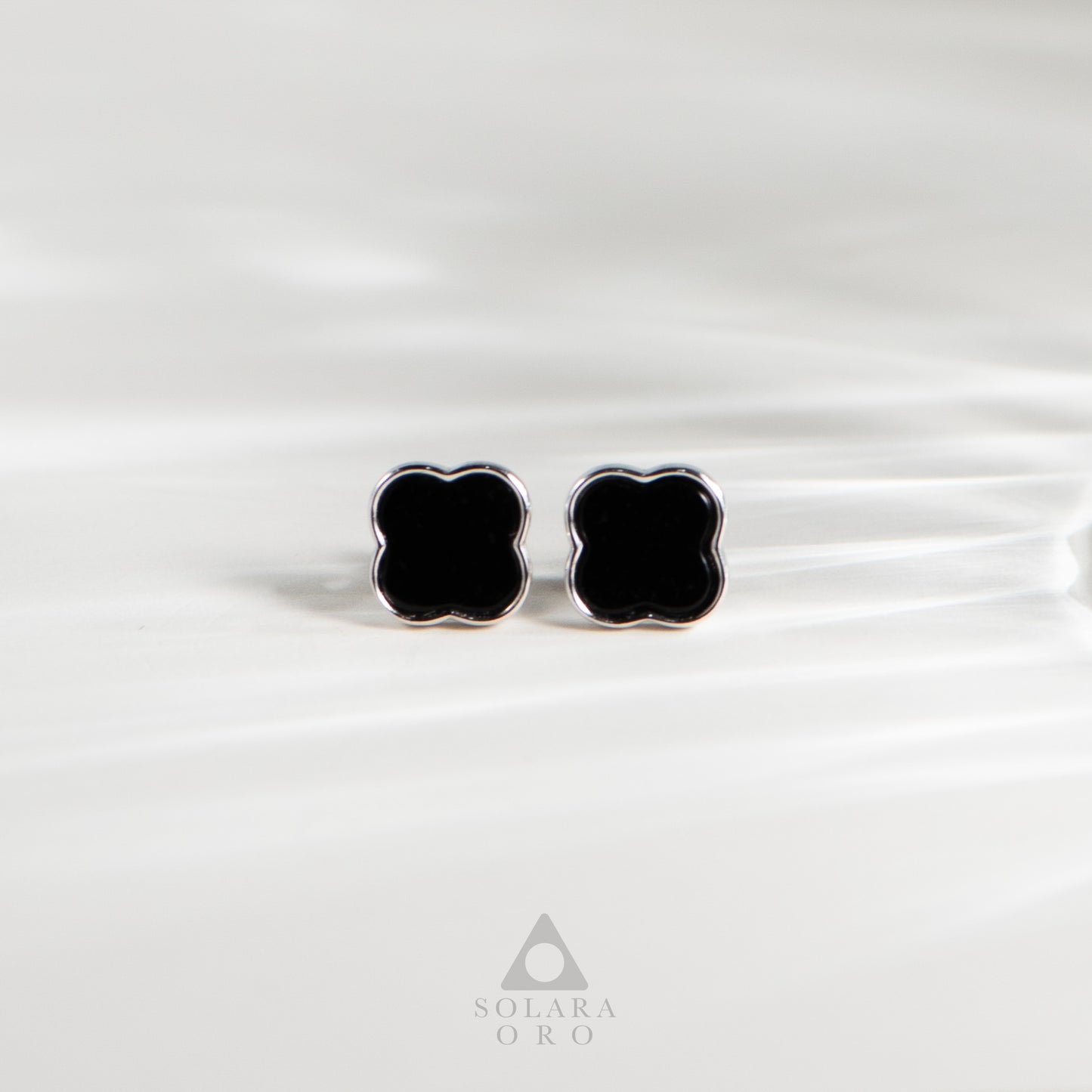 Black clover-shaped earrings on a white satin background with Solara Oro logo.