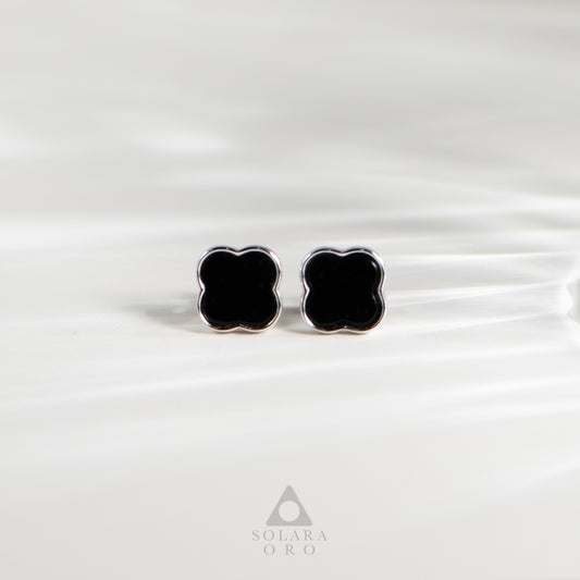 Black clover-shaped earrings on a white satin background with Solara Oro logo.