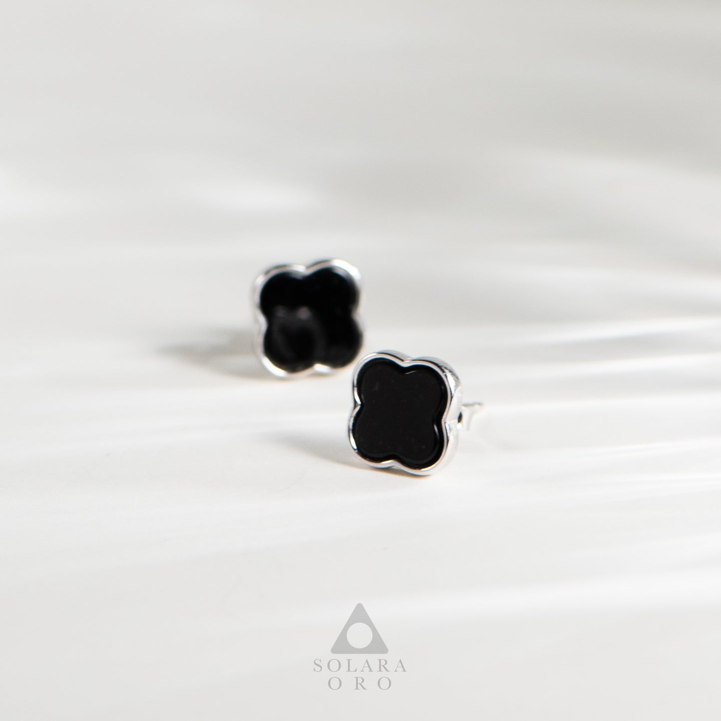 Black clover-shaped earrings on a white satin background with 'Soleara Oro' branding.