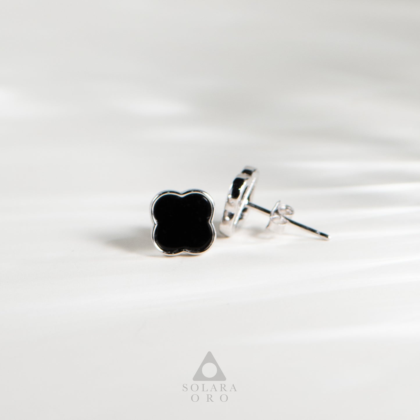 Black clover-shaped earrings on a white background with 'Soleara Oro' branding.