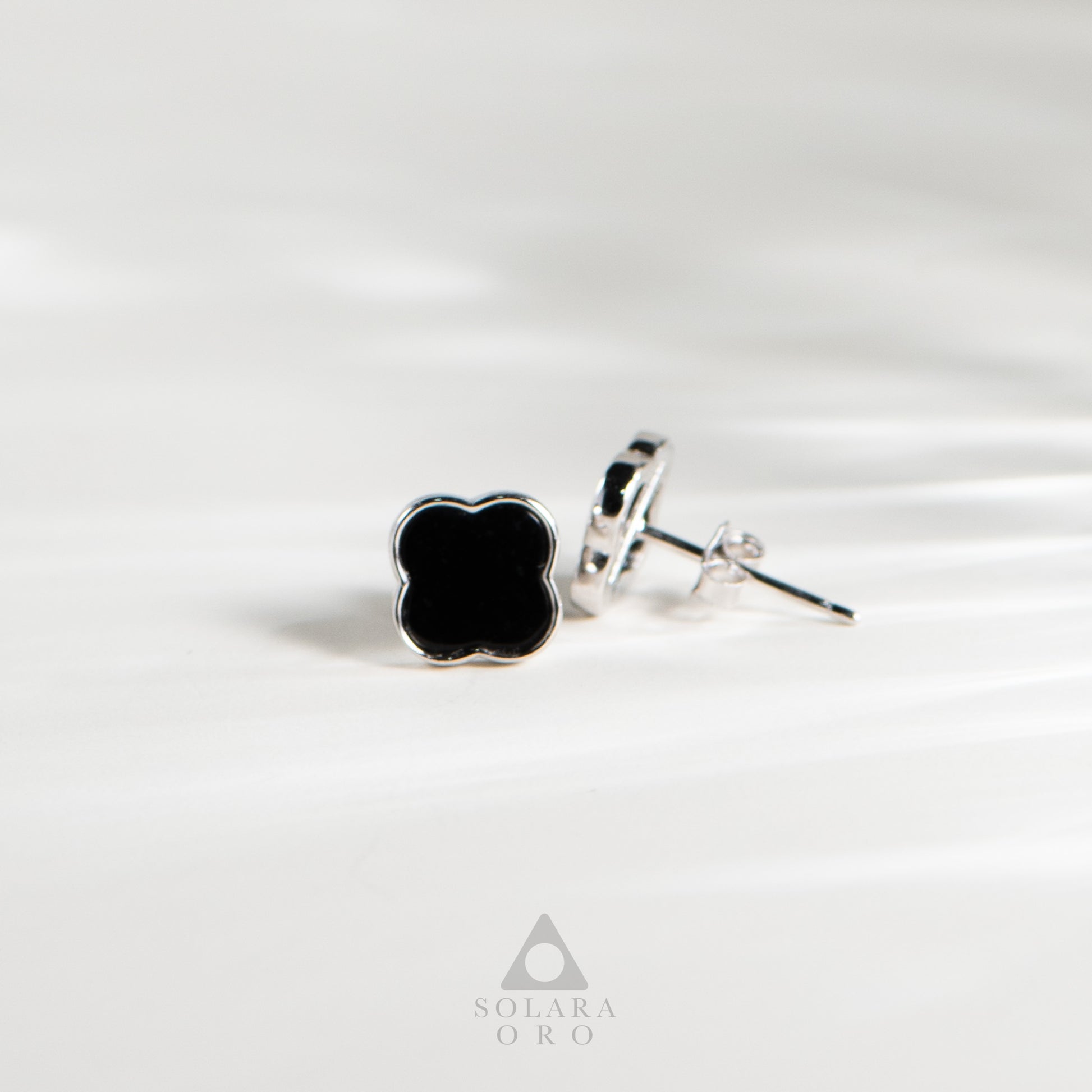 Black clover-shaped earrings on a white background with 'Soleara Oro' branding.