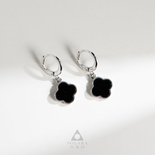 Pair of black flower-shaped earrings with silver hooks on a light gray background, featuring the Solara Oro brand.