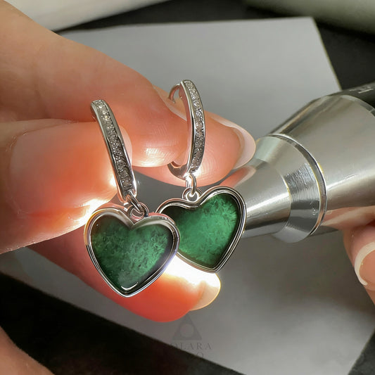 Silver heart-shaped earrings with green stones held by a hand against a neutral background