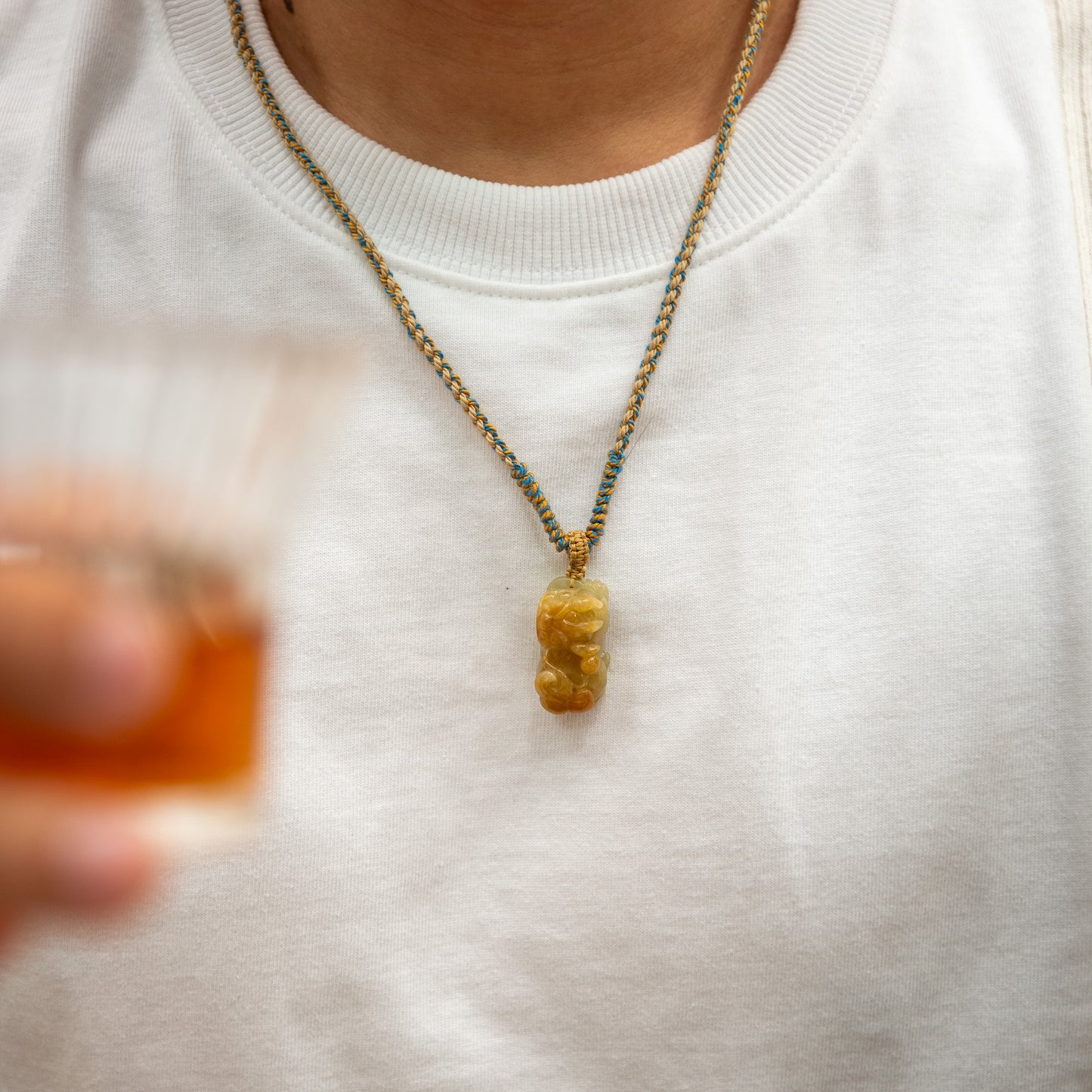 Person wearing a necklace with a bear pendant on a white shirt, holding a blurred drink.