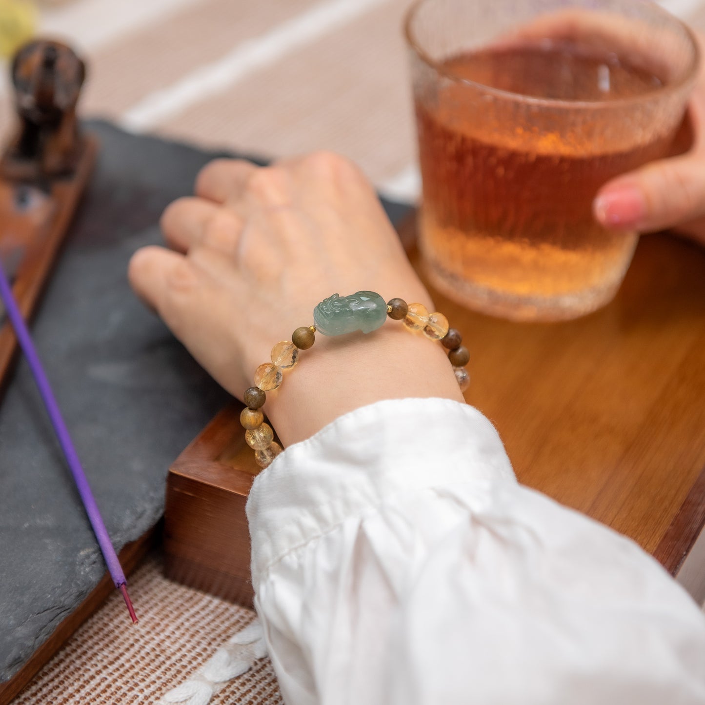 Person wearing a beaded bracelet with a green stone, holding a glass of amber liquid on a wooden surface.