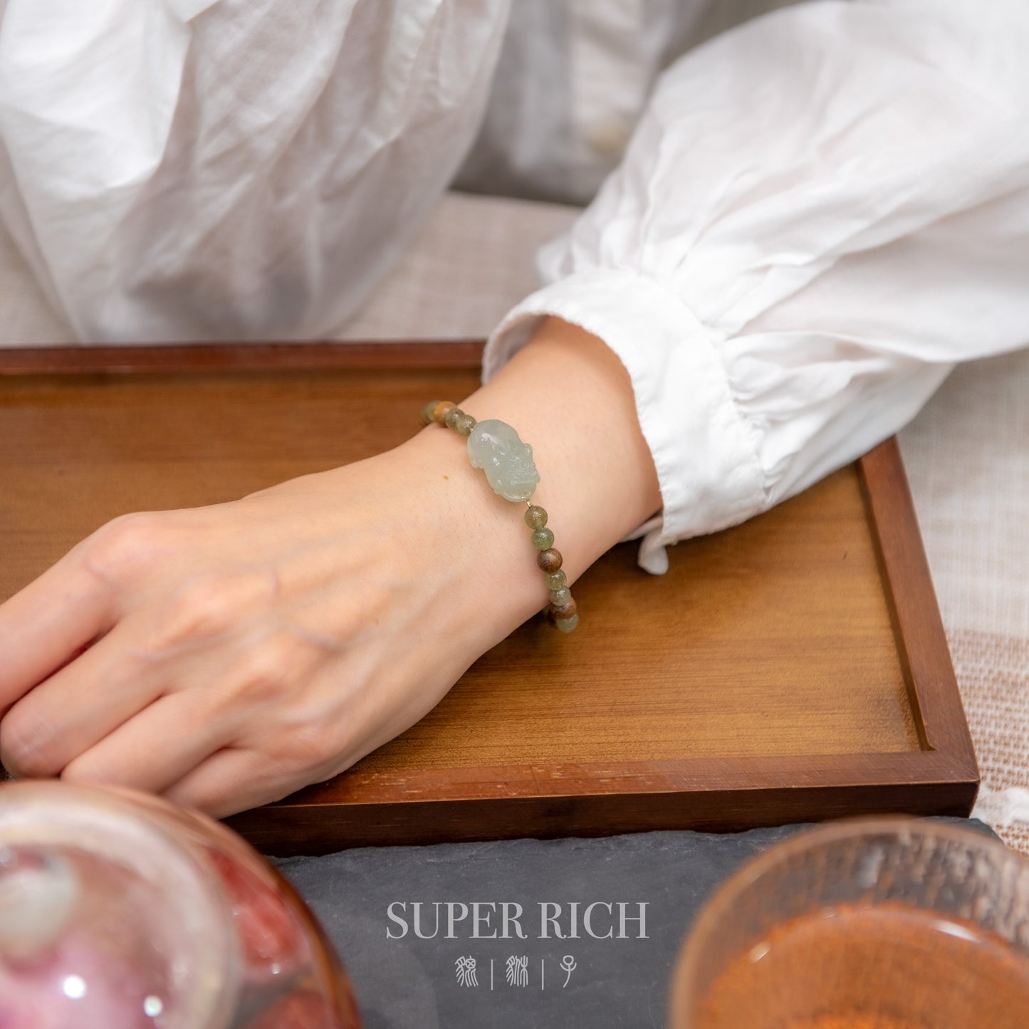 Hand wearing a bracelet on a wooden tray with 'Super Rich' branding.