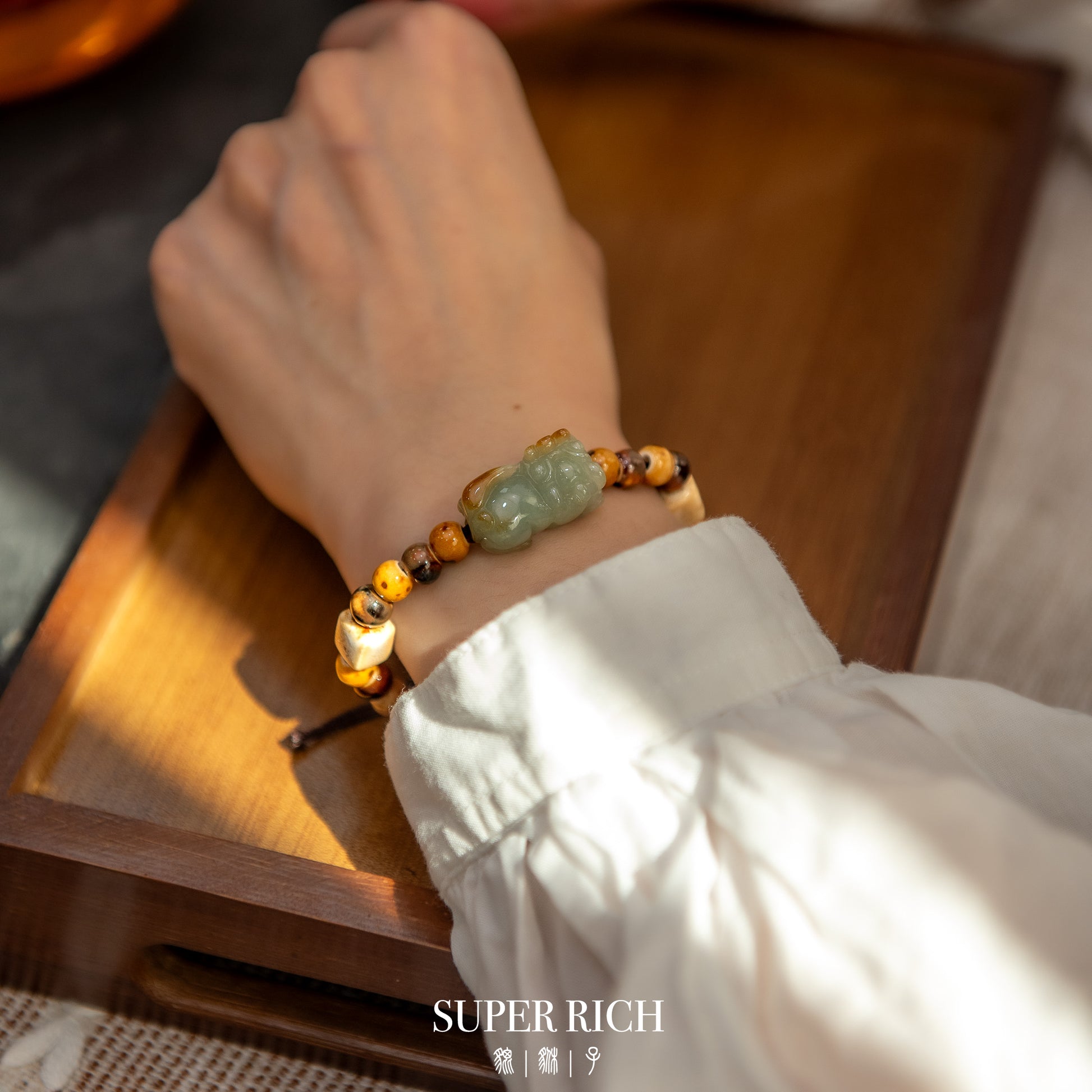 Hand wearing a beaded bracelet with a green stone on a wooden surface, 'SUPER RICH' brand visible.