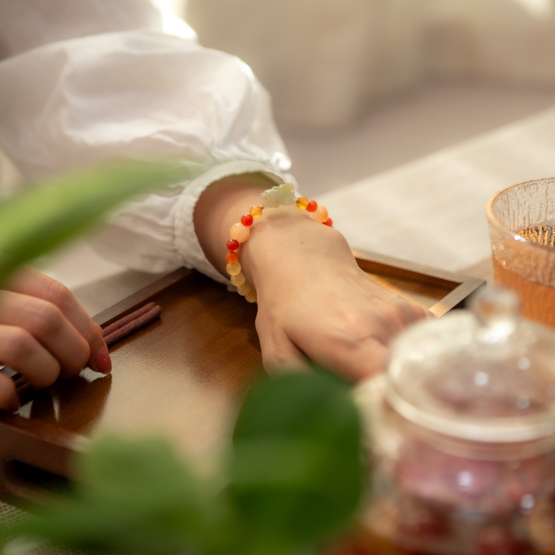 Person wearing a colorful beaded bracelet on a wooden table with a plant and teapot in the foreground.